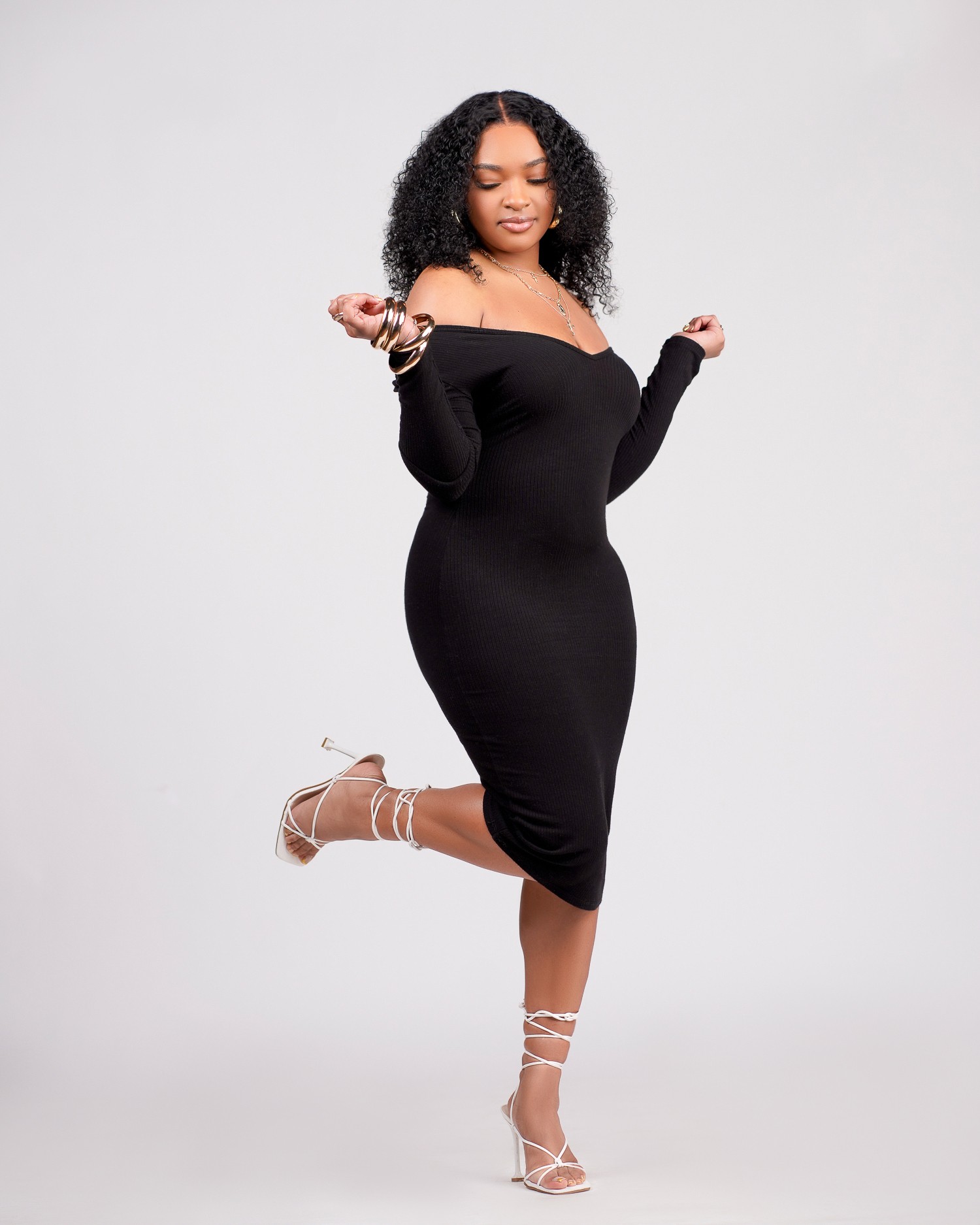 Woman in black off-shoulder dress and high heels poses with one leg raised and arms bent, against a plain background.