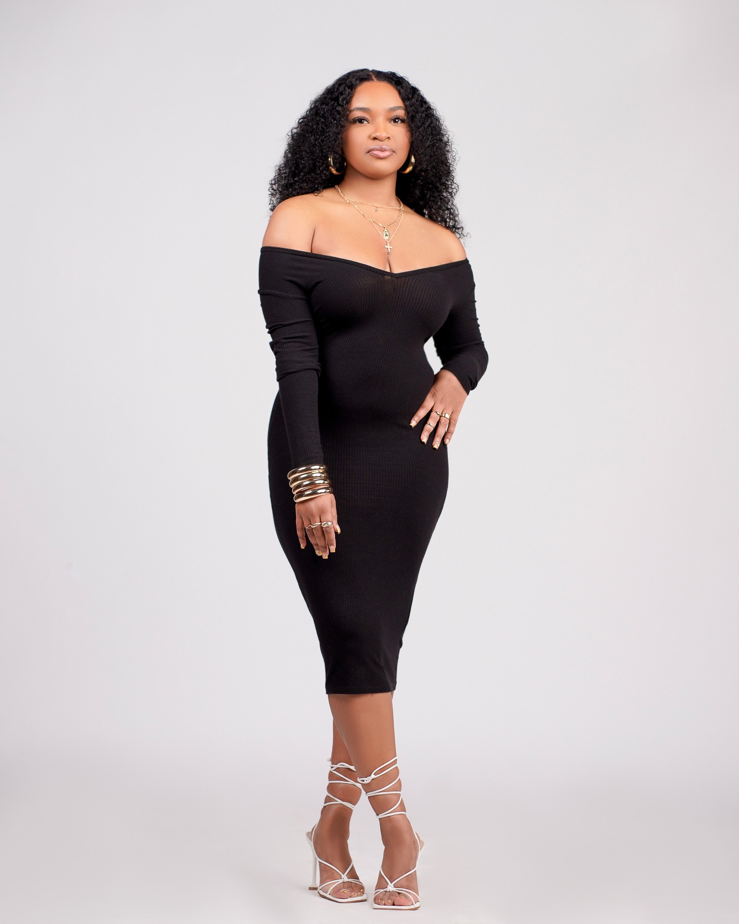 Woman in black off-shoulder dress with jewelry and strappy heels, standing against a plain background.