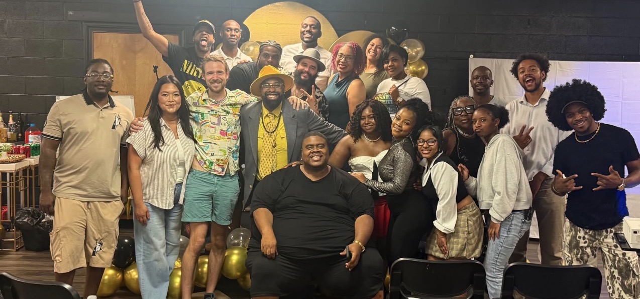 Group of people posing together indoors, some smiling, with a yellow backdrop and balloons, diverse in age and appearance.