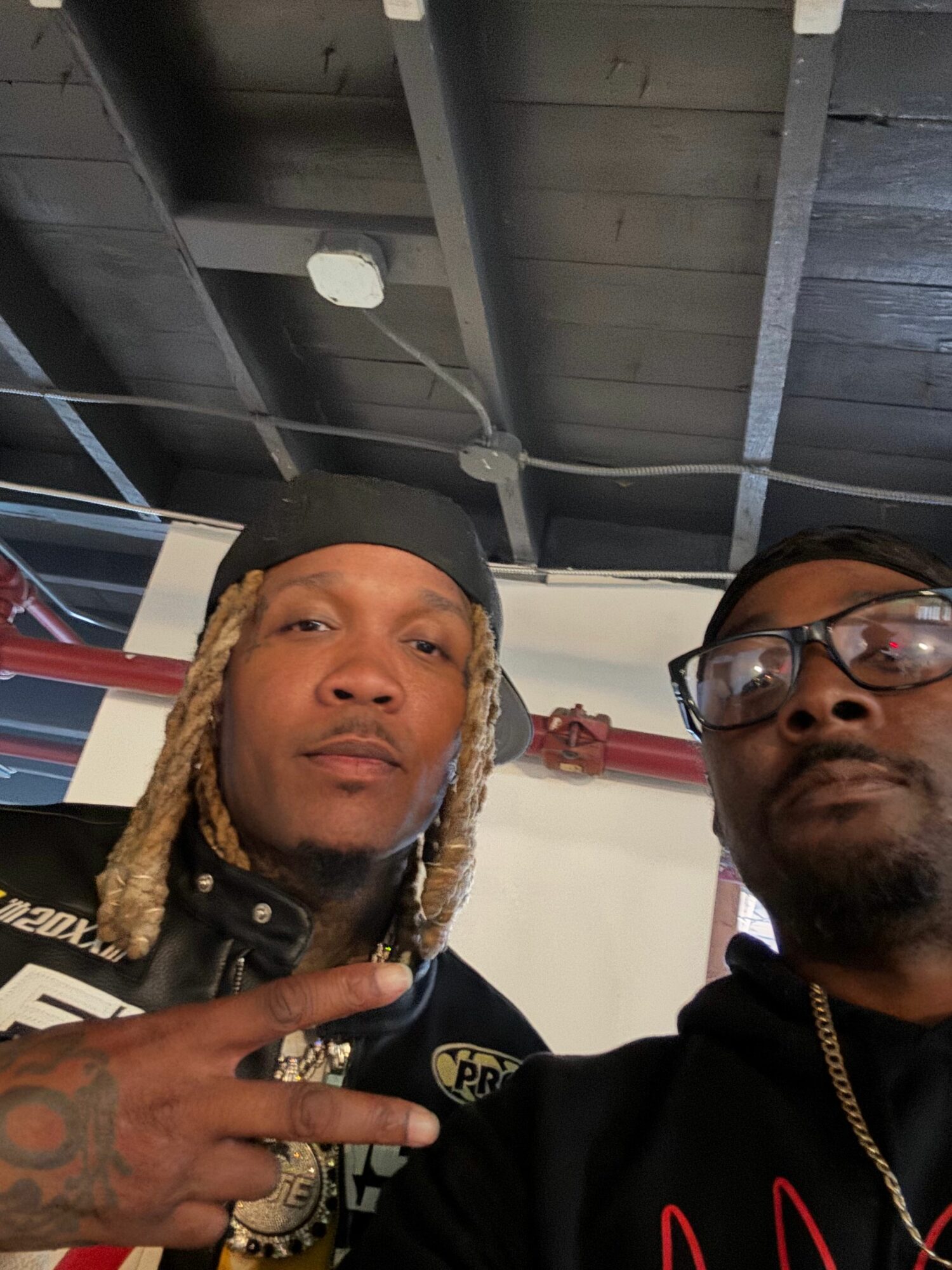 Two men pose indoors, one making a peace sign, both looking at the camera.