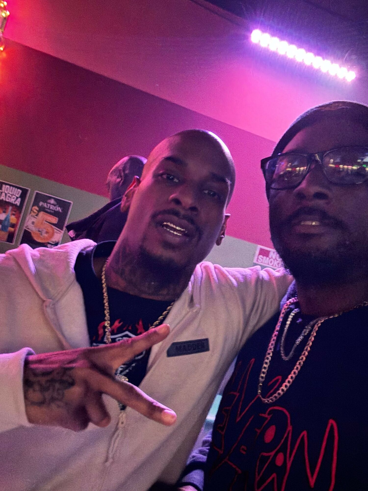 Two men smiling, one making a peace sign, in a room with purple lighting.
