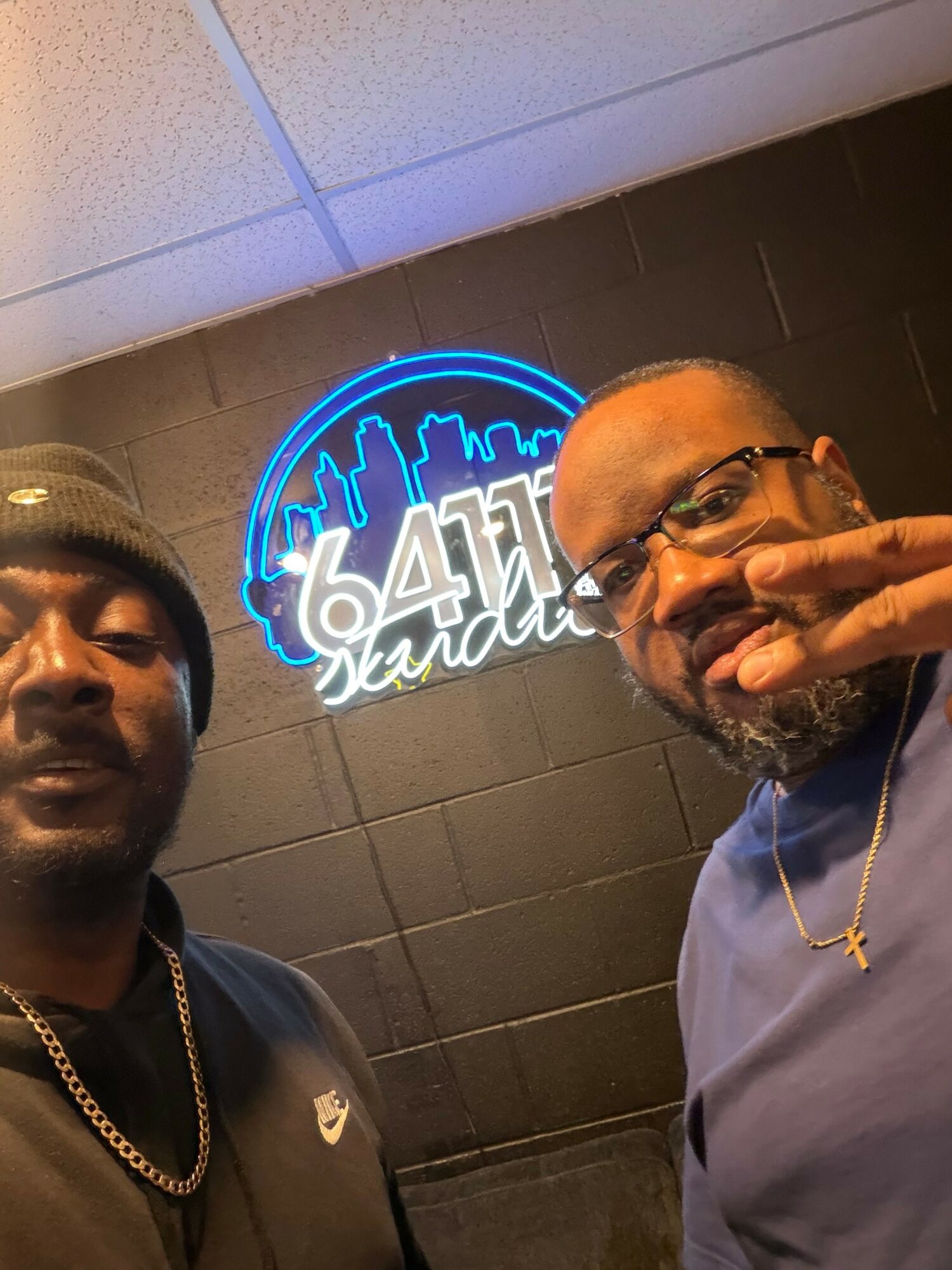 Two men taking a selfie in front of a black wall with a neon blue city skyline and text reading '6411 Sandtown'.
