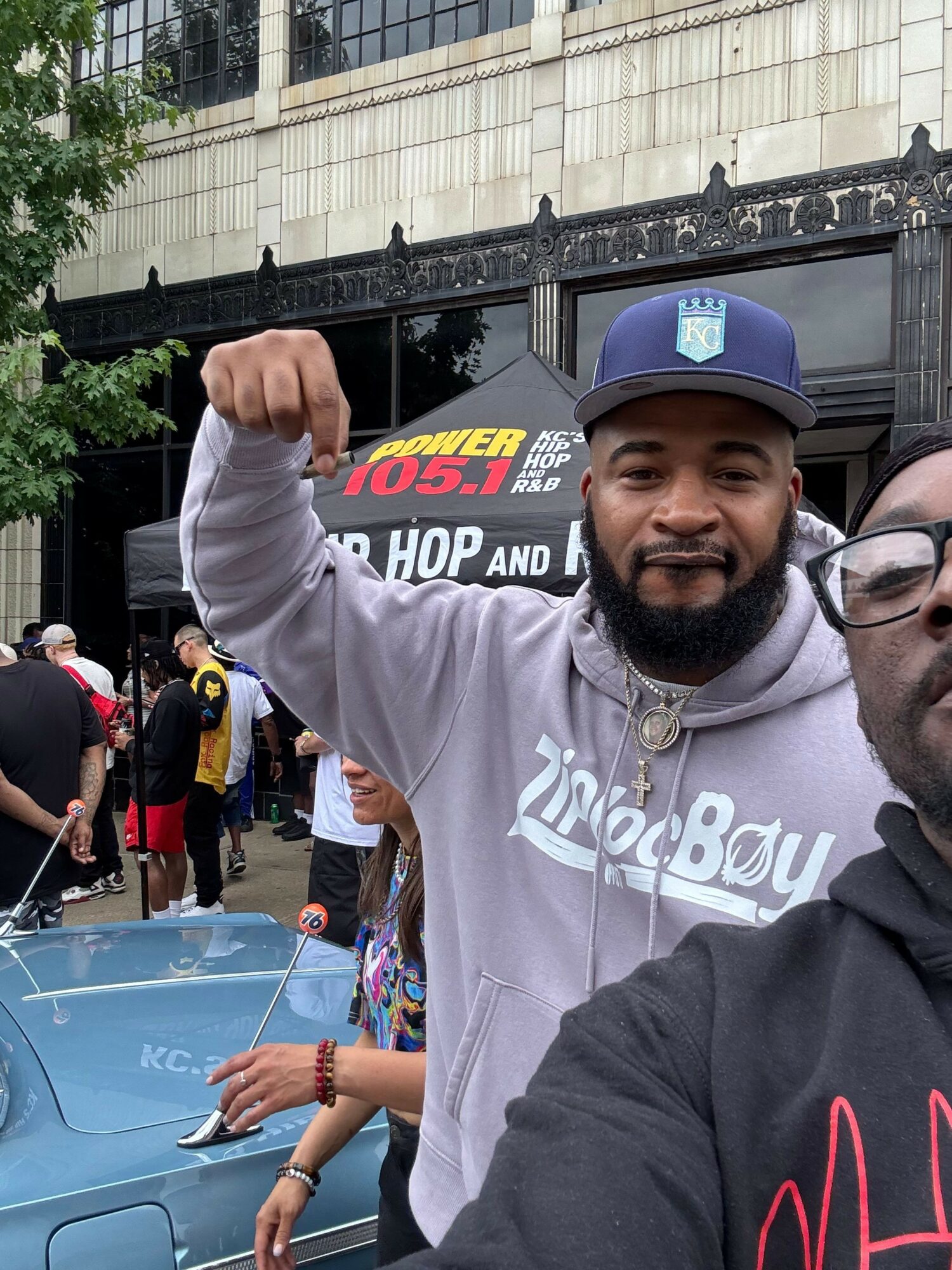 Two men taking a selfie outdoors, one wearing a baseball cap and hoodie, the other partially visible, with a building and people in the background.