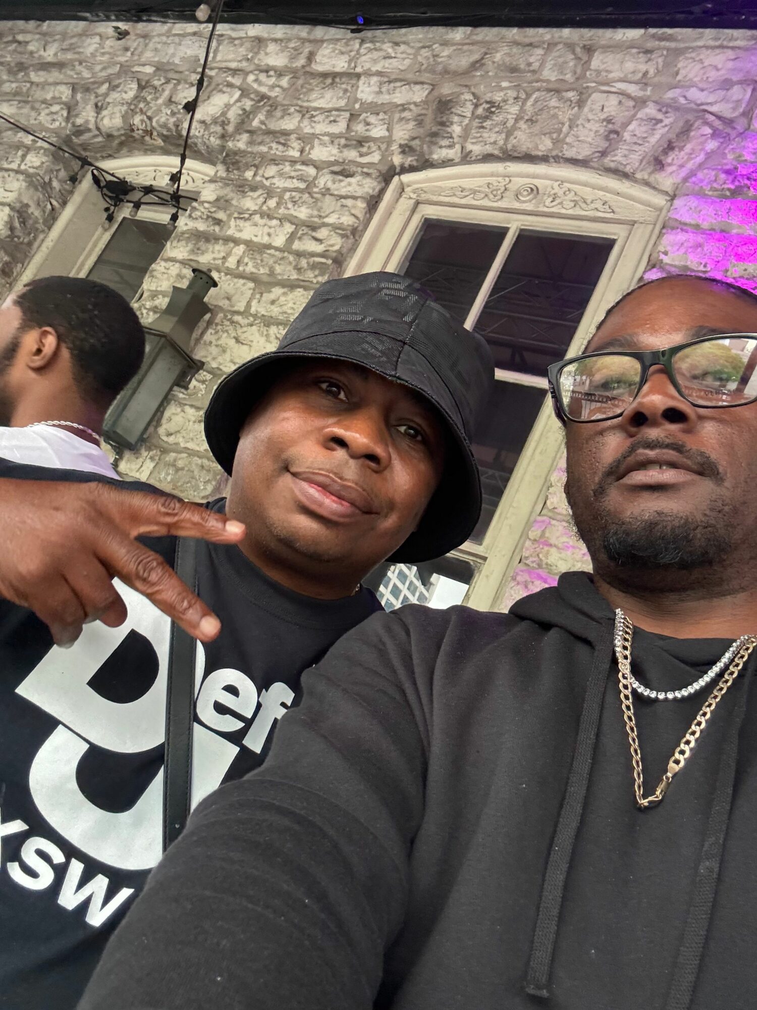Two men taking a selfie outdoors with a brick building and window in the background.