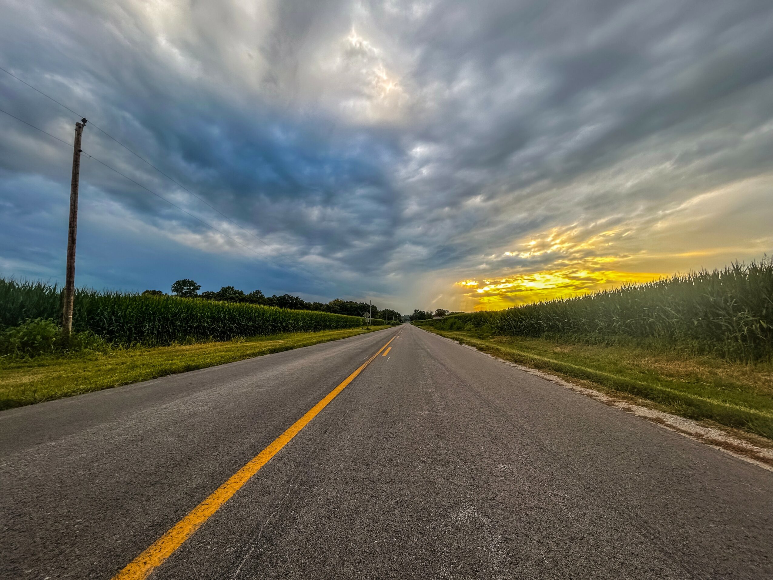 Straight road with yellow dividing line, cloudy sky, and sunset in the distance, flanked by green fields and trees.