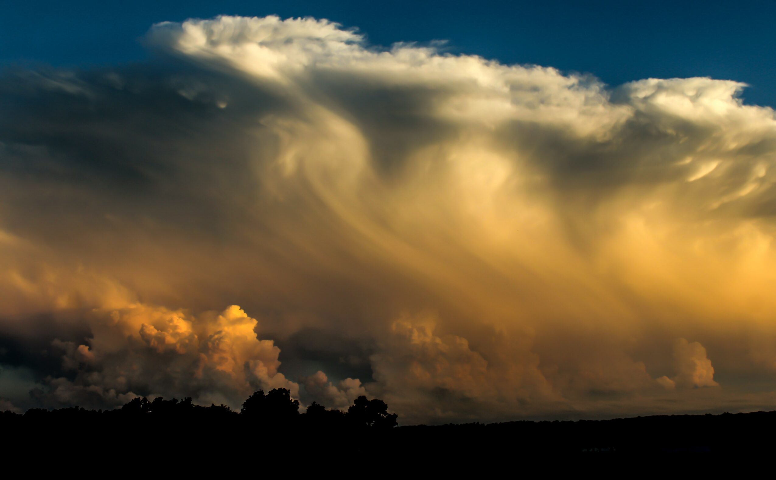 Cloudy sky with dark and light clouds during sunset or sunrise, with a silhouette of trees or landscape at the bottom.