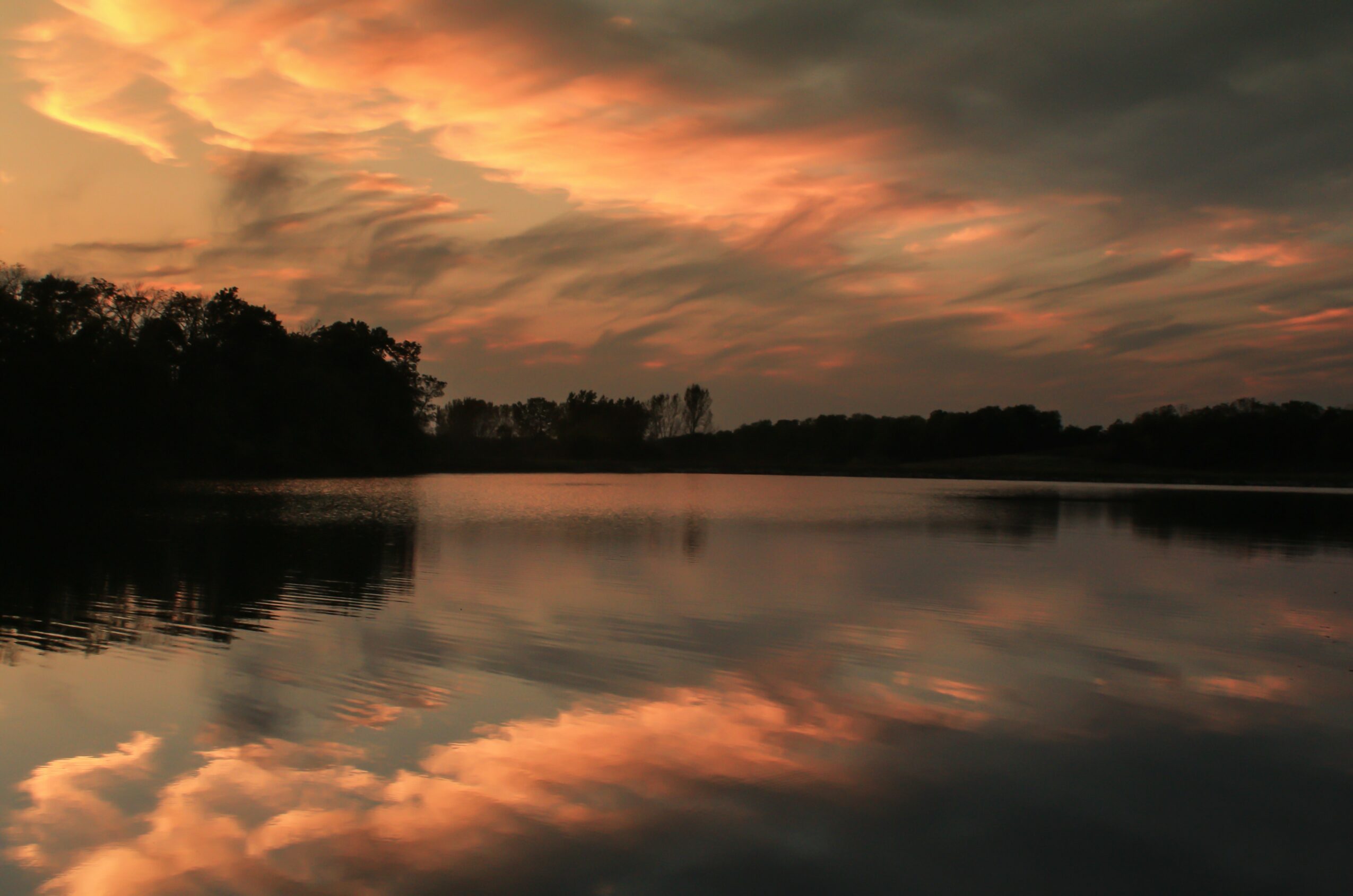 Sunset sky with orange and pink clouds reflected on a calm river, with trees along the horizon.