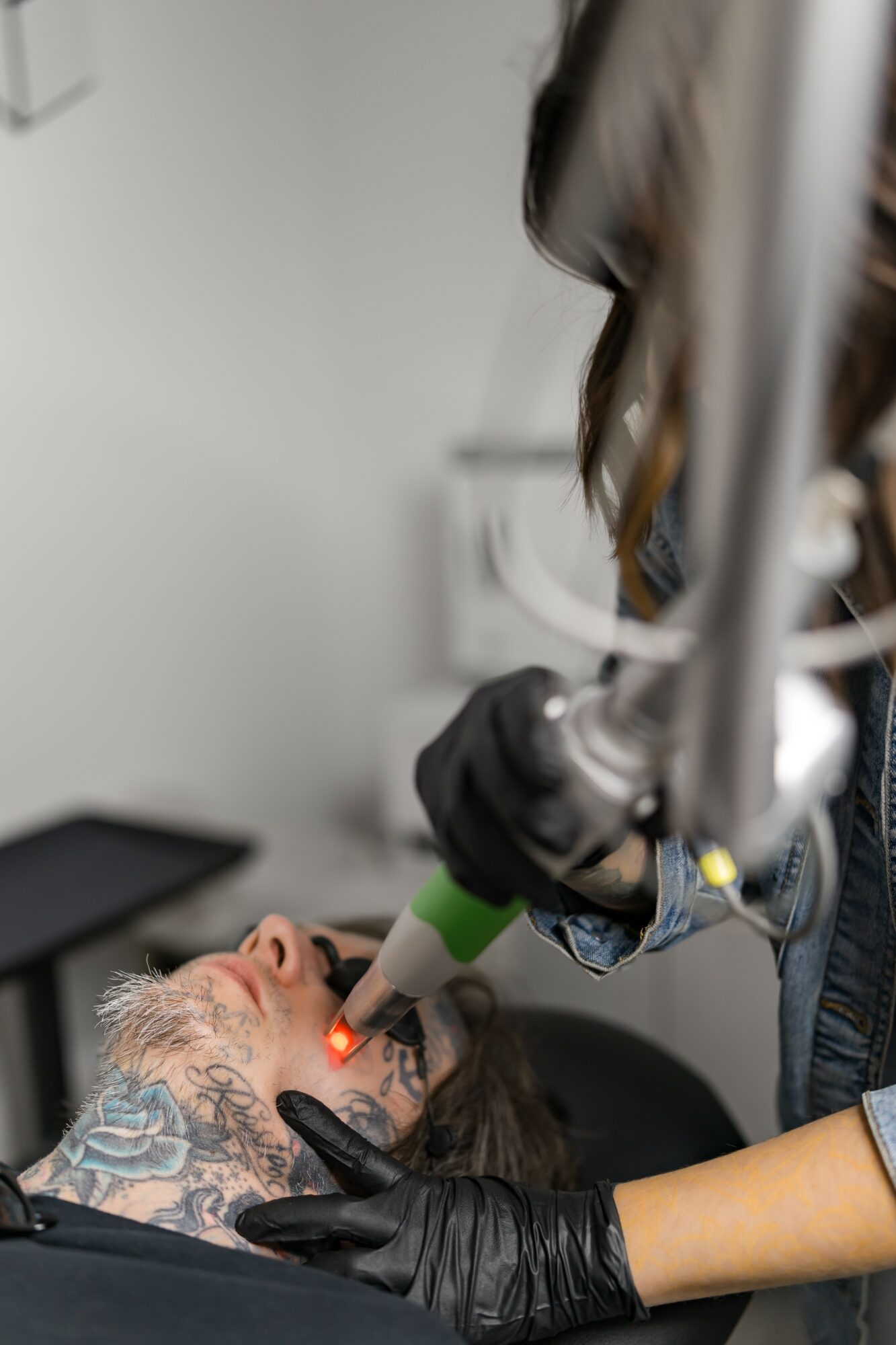 Person receiving a tattoo on face, lying down with head tilted back, tattoo artist using a tattoo machine, wearing black gloves.