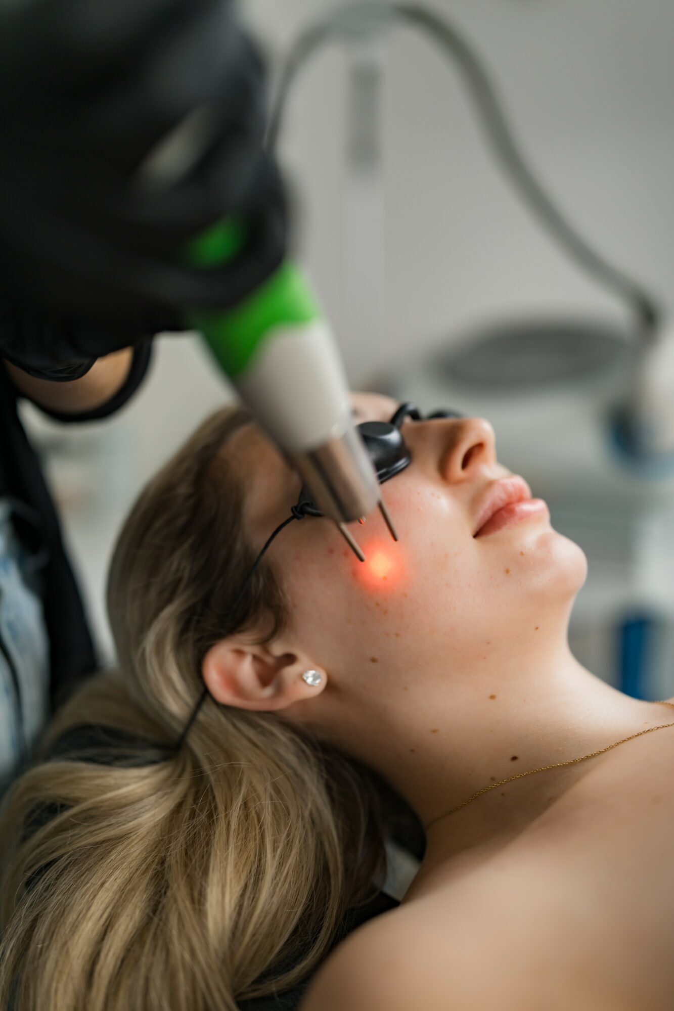 Woman receiving laser eye treatment with a device emitting a red light, lying down with eyes closed, in a clinical setting.