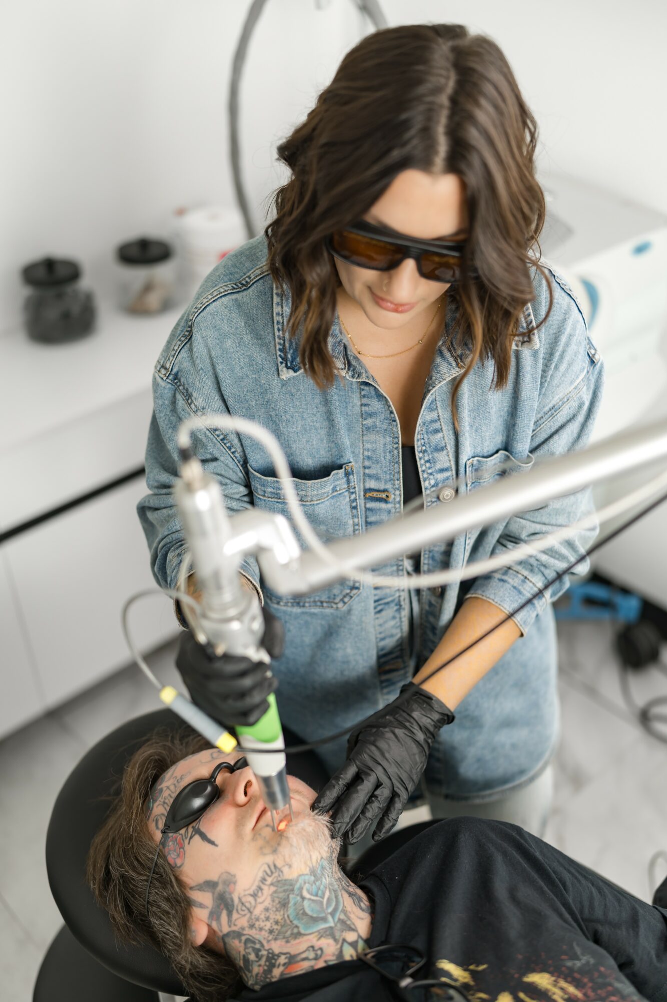 Woman in sunglasses and denim jacket operating a medical or cosmetic device on a person with tattoos.