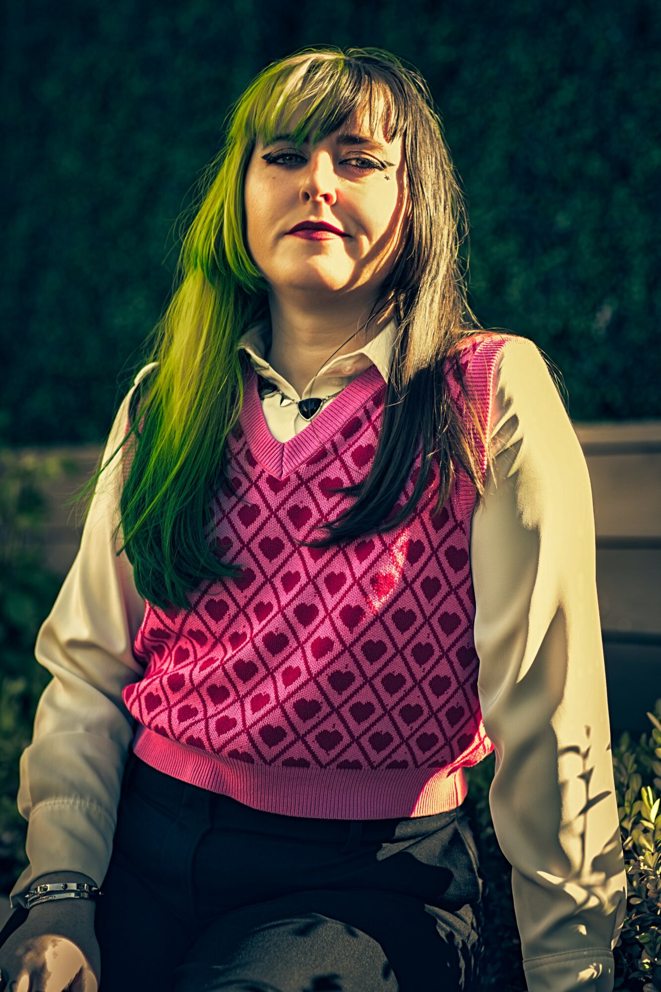 Young woman with long hair, wearing a pink patterned vest over a cream blouse, standing outdoors at night.