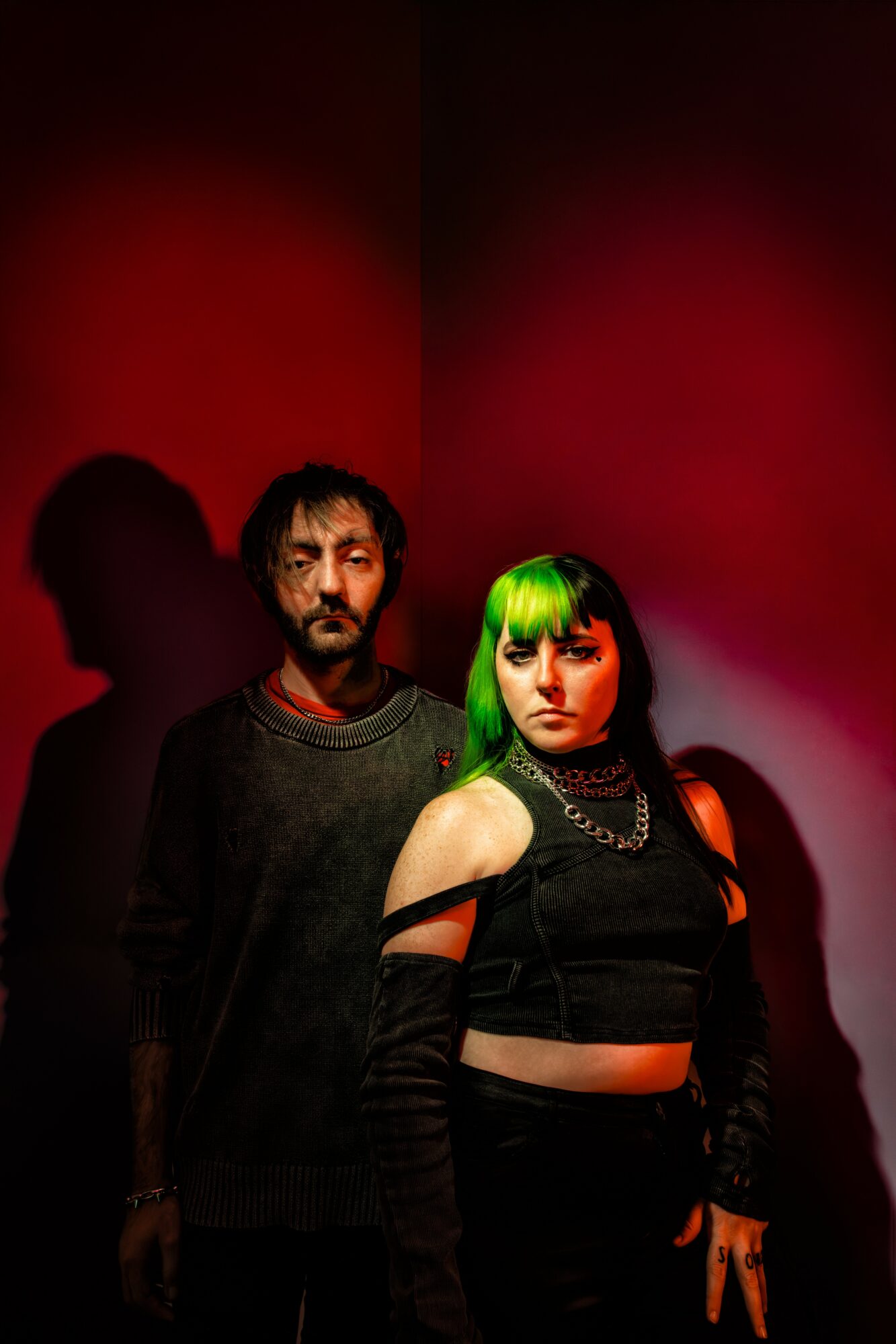Two people standing against a red background, one with green hair and the other with dark hair, both dressed in dark clothing.