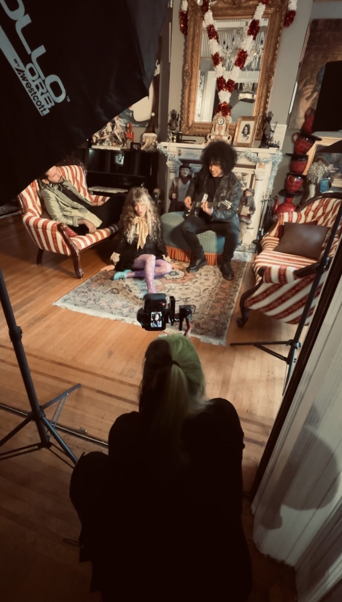Two people sit on a patterned rug in a decorated room with chairs, a mirror, and various objects, with a camera on a tripod in front.