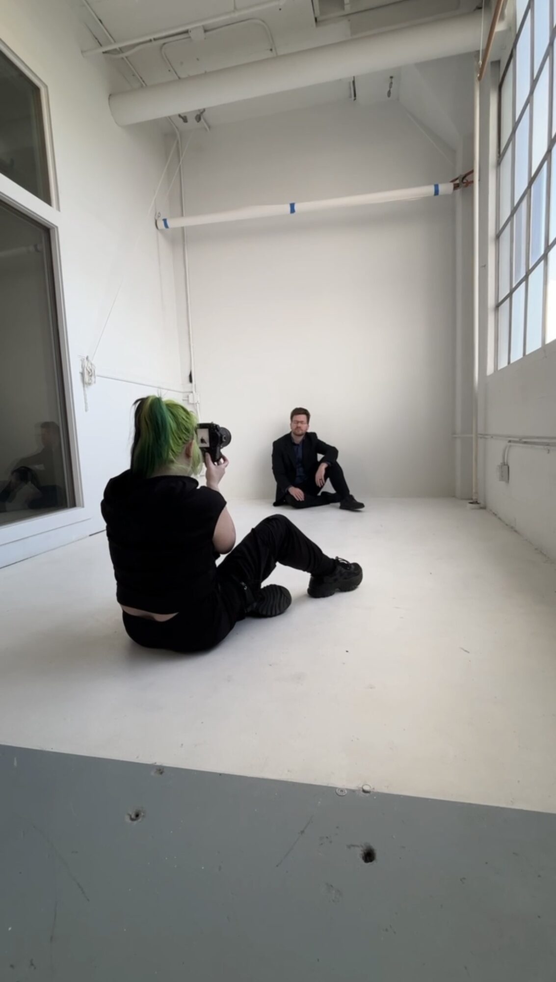 Two people sitting on a white floor in a bright room with large windows, one with green hair and the other with dark hair.