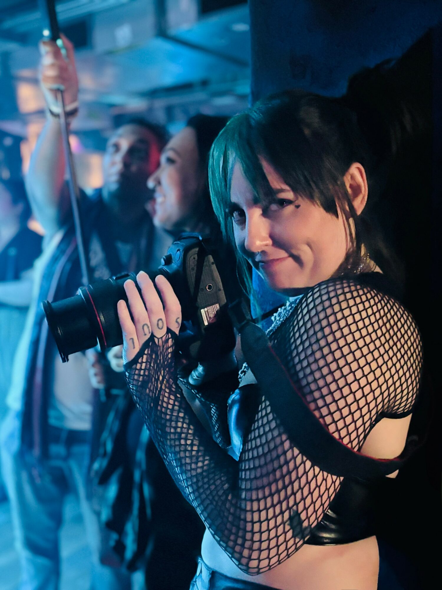 Young woman with black hair and fishnet top holding a microphone, smiling in a club setting with blue lighting.