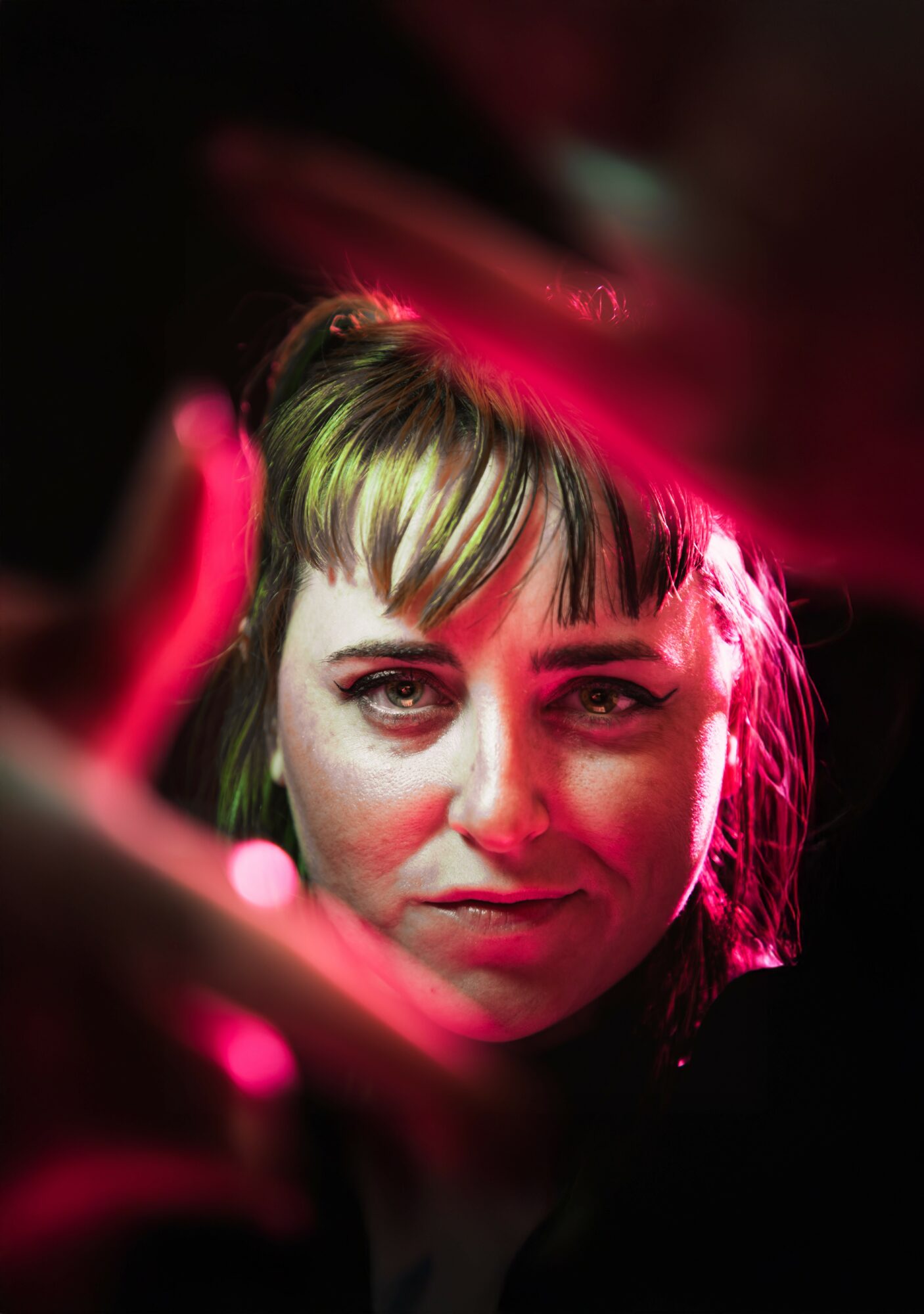 Close-up of a woman with short hair and makeup, illuminated by pink and red lighting, looking at the camera.