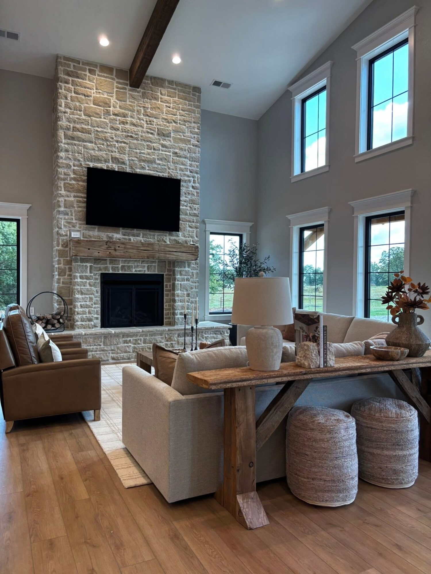Living room with a brick fireplace, wall-mounted TV, large windows, and seating area with sofas and stools.