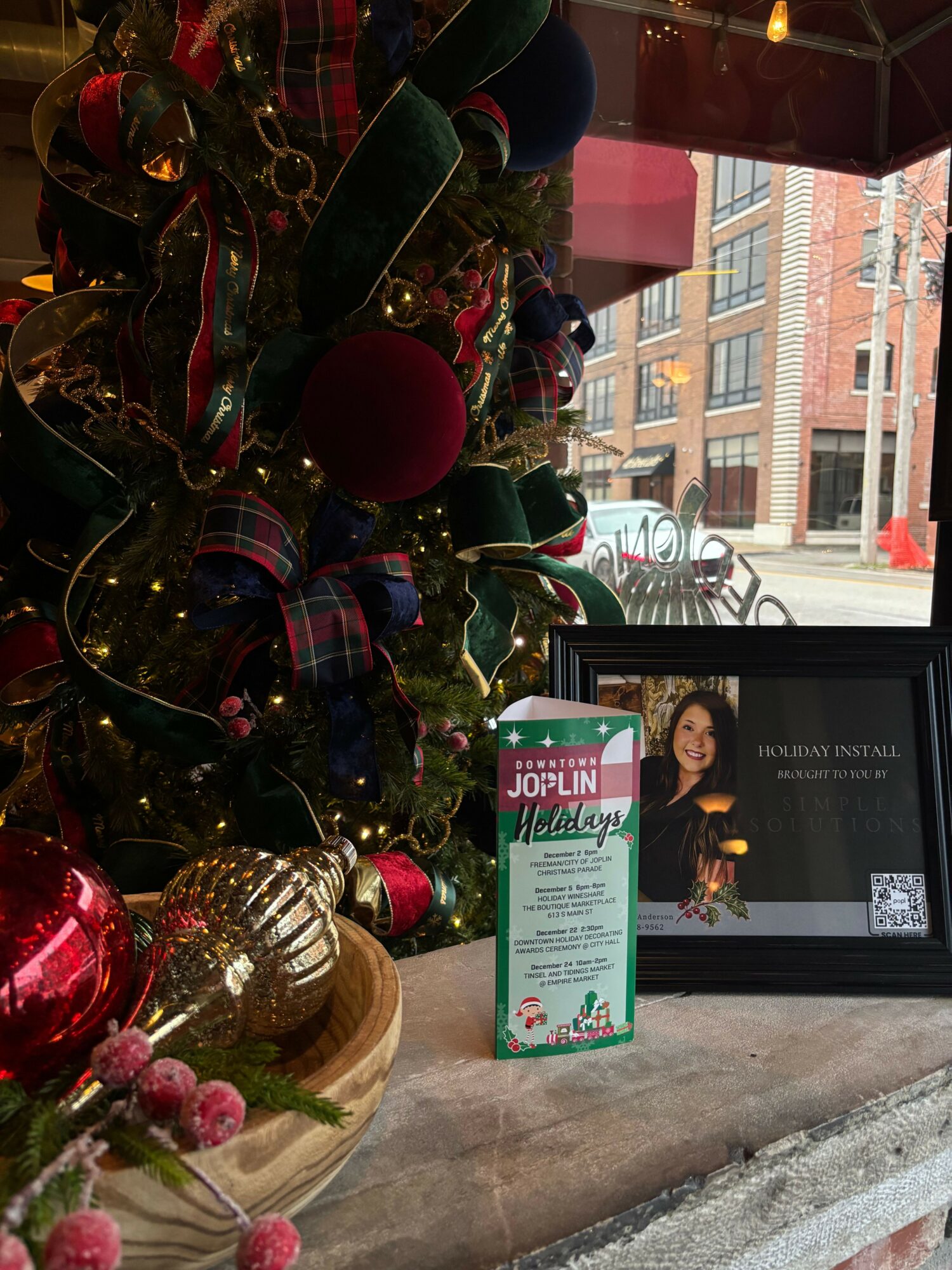 Decorative Christmas ornaments and ribbons near a window with a framed photo and a sign, outside city buildings visible.