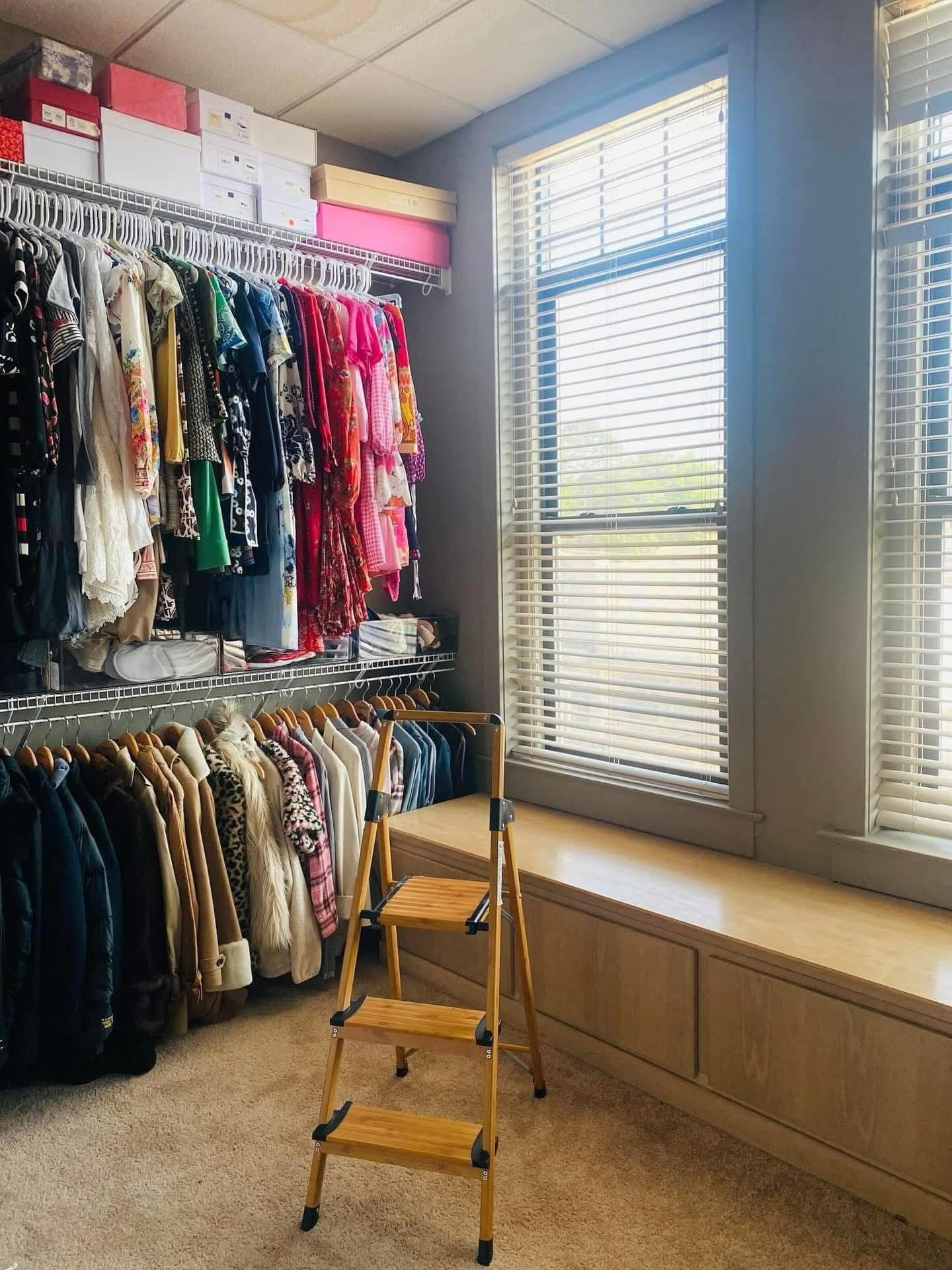 Closet with hanging clothes, window with blinds, and a small wooden step ladder, in a well-lit room.