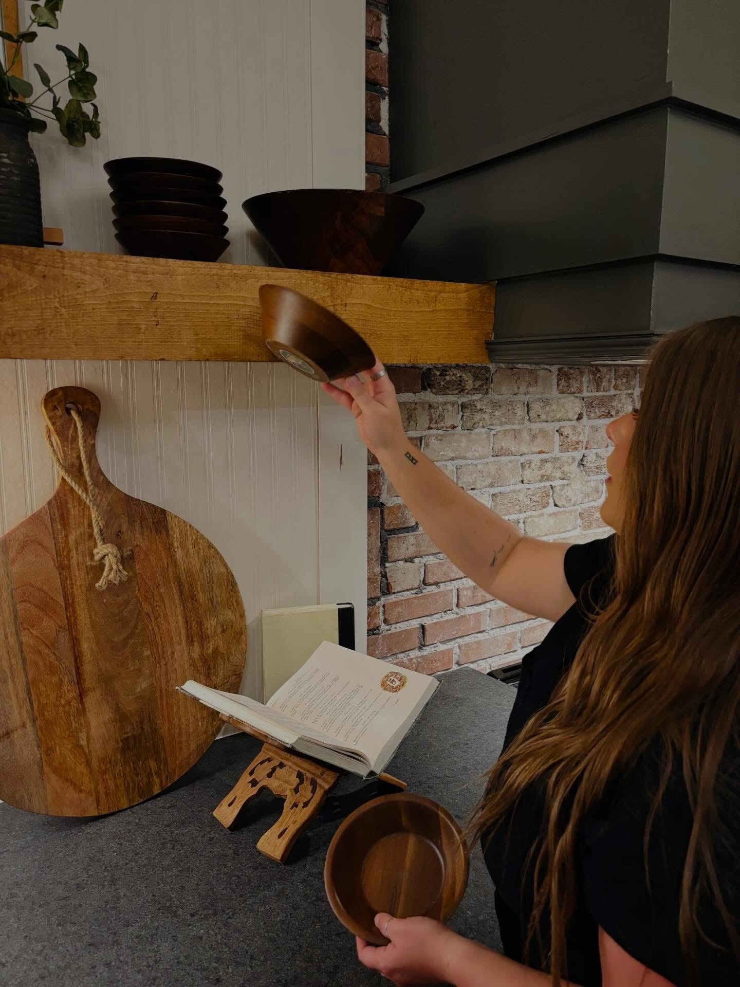 Woman reaching for a bowl on a wooden shelf, with a book and cutting board nearby, brick wall in background.