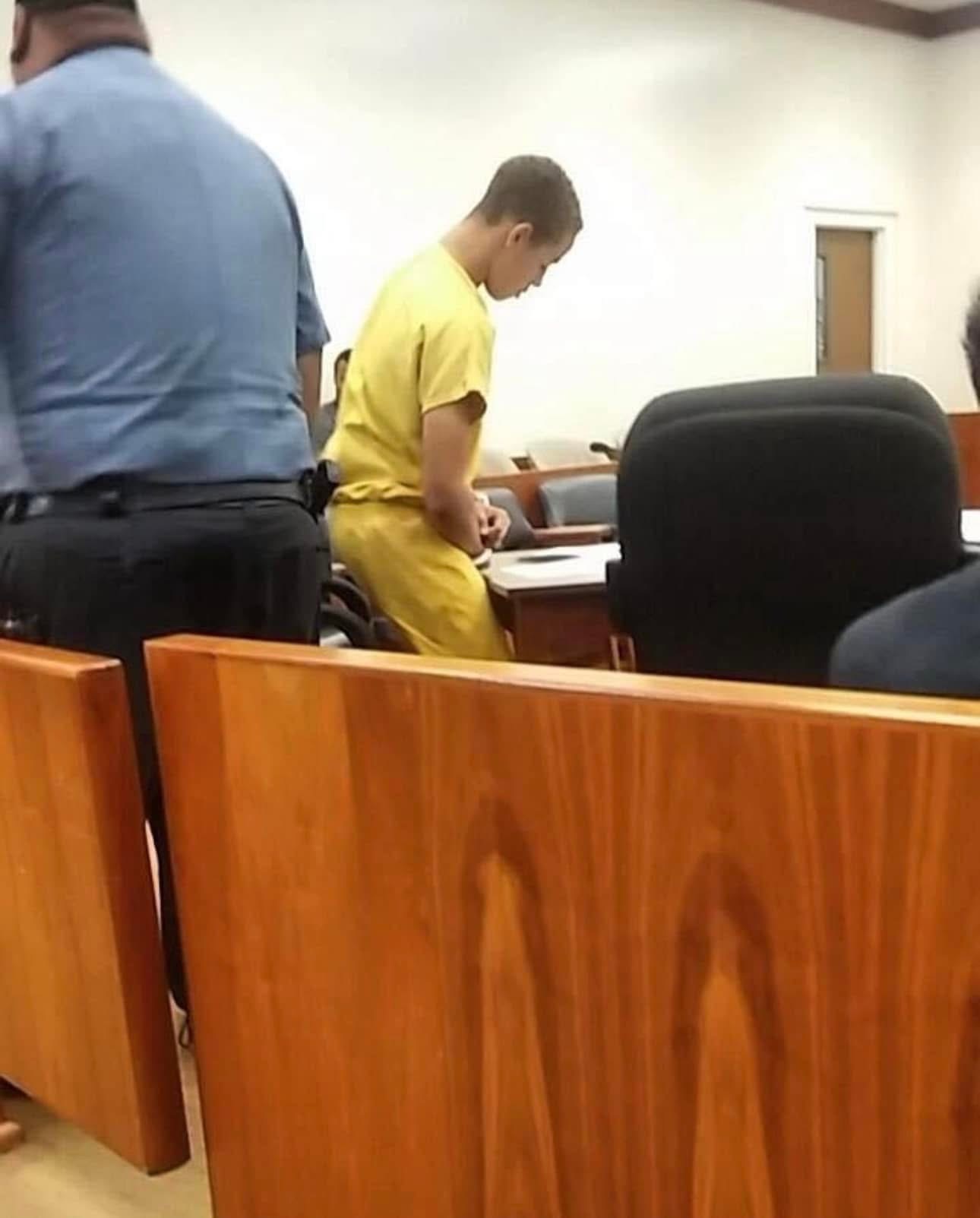Young person in yellow clothing standing in a room with chairs and wooden furniture, facing downward, with other people nearby.