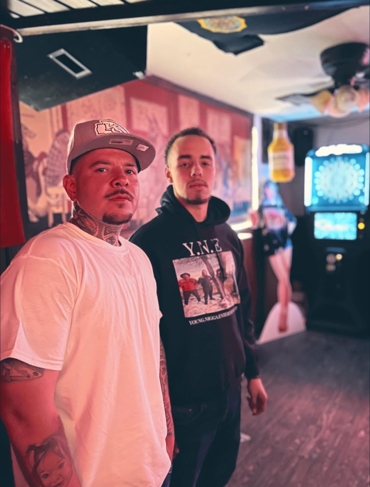 Two men standing in a dimly lit arcade or gaming area with a colorful background and arcade machines.