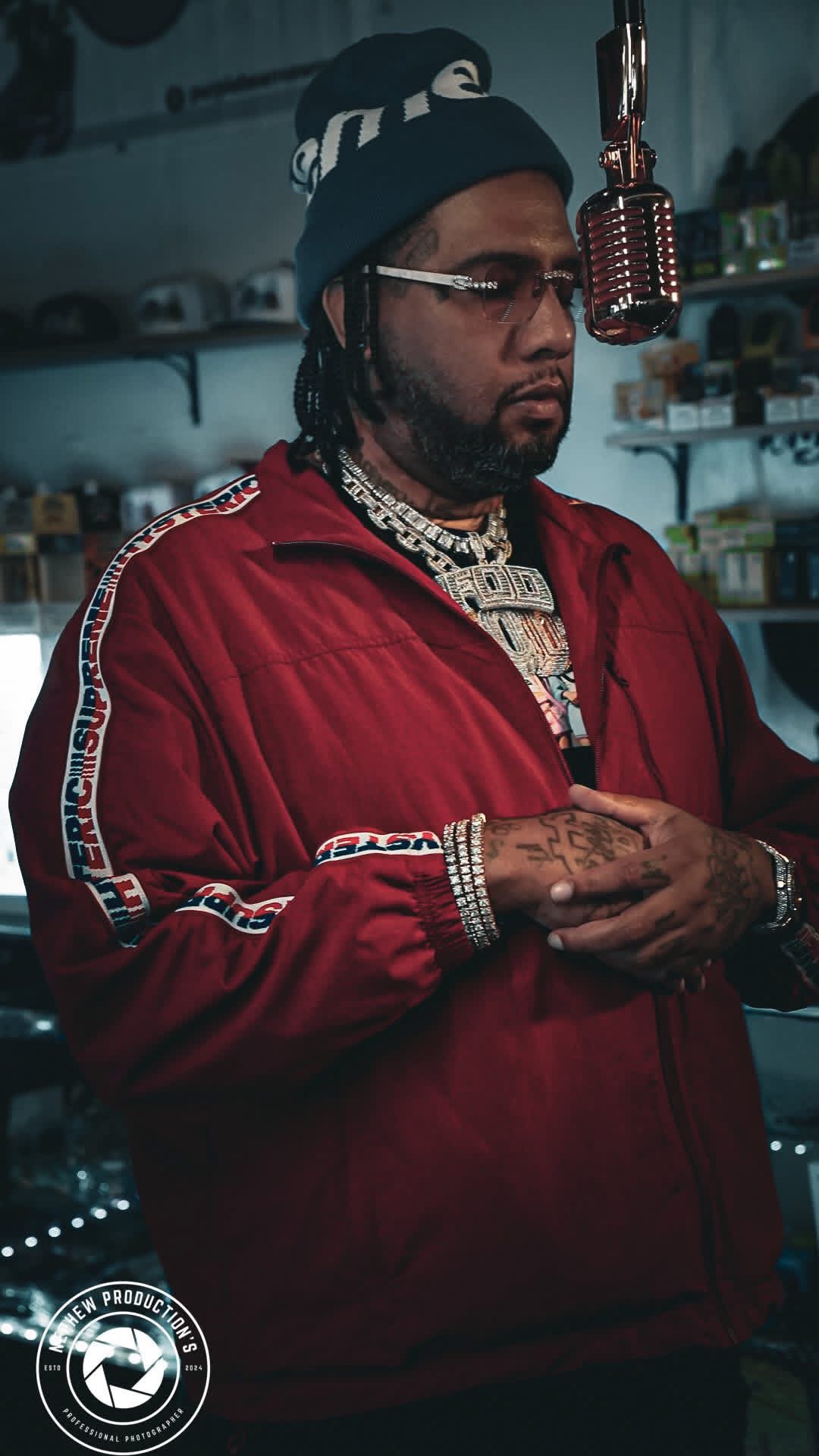 Man with glasses and jewelry in a store, wearing a red jacket and a beanie, standing near a microphone.