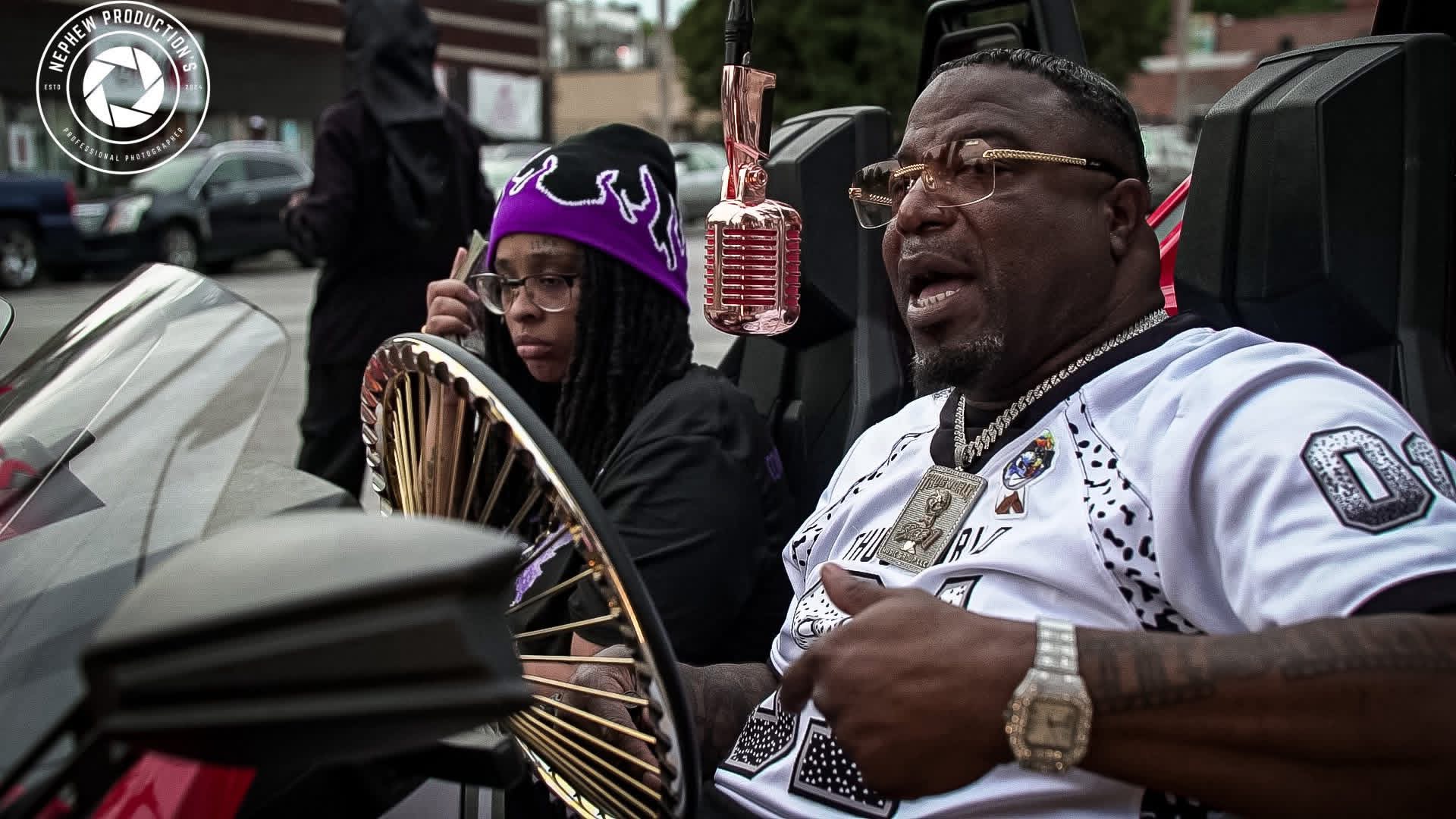 Two people sitting in a vehicle, one wearing a purple crown hat and the other speaking, with a microphone and fan visible.