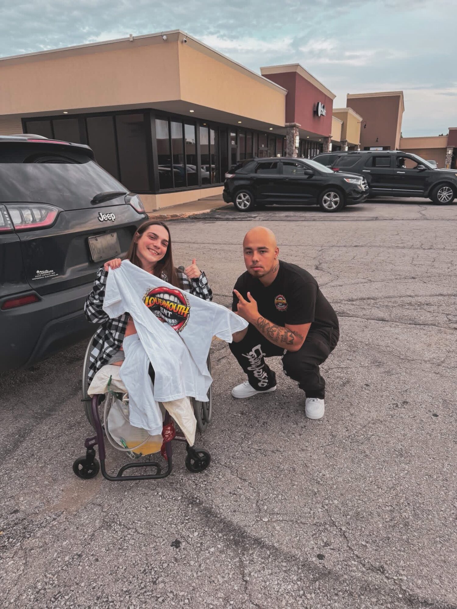 A woman sitting in a shopping cart holding a white T-shirt with a colorful design, with a man crouching beside her in a parking lot.