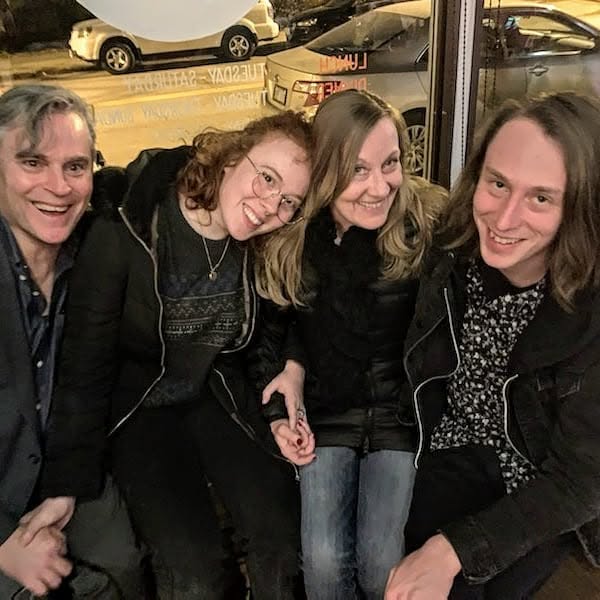 Four people smiling and sitting together indoors, with cars visible outside through the window behind them.