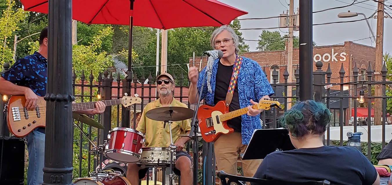Group of musicians performing outdoors under a red umbrella, with a woman playing guitar and singing, and a drummer nearby.