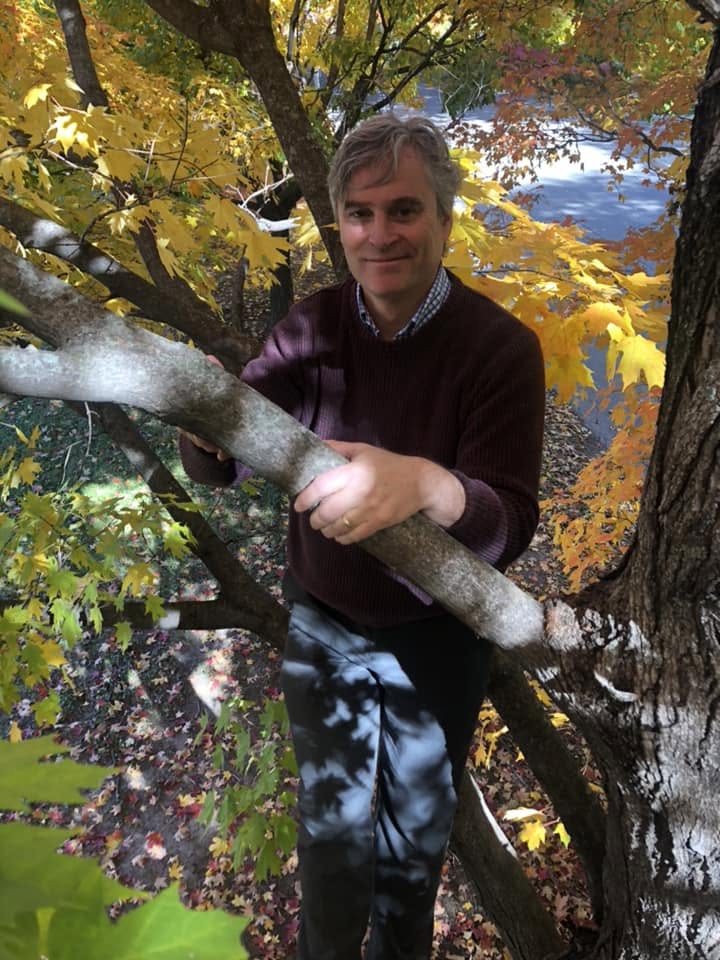 Man standing outdoors among autumn trees with yellow and green leaves, holding a snow-covered branch, smiling at the camera.