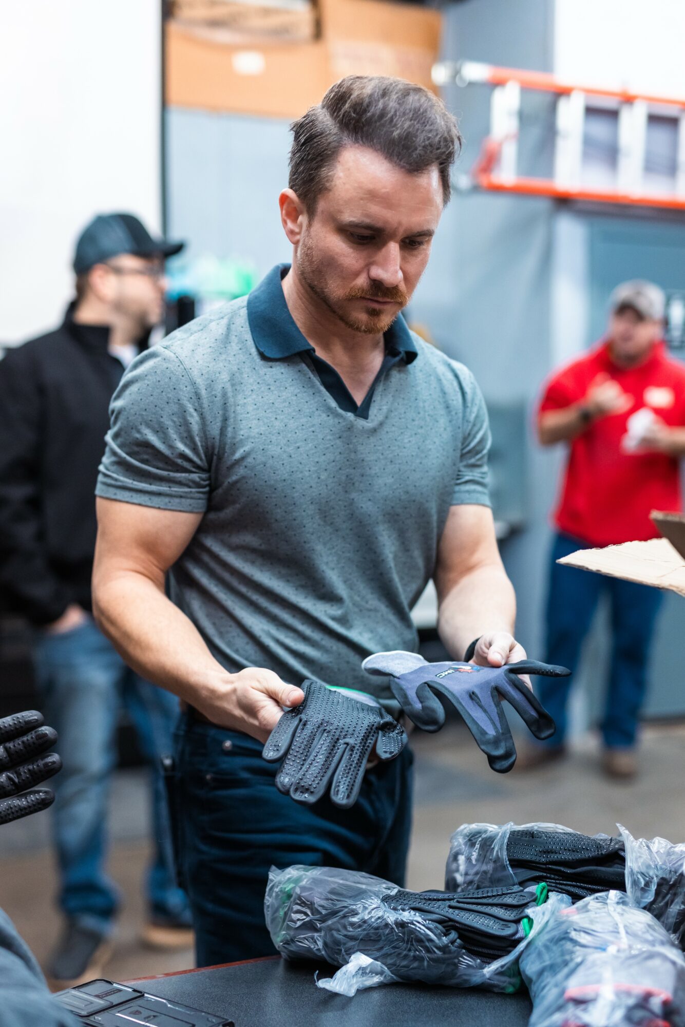 Man in gray polo shirt holding gloves at a table with others in background, workshop setting