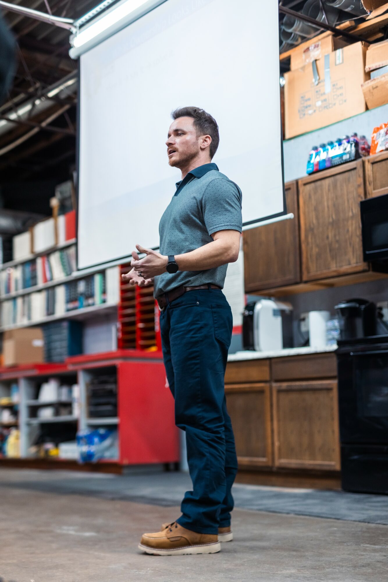 Man standing and speaking in front of a large screen in a warehouse or storage area with shelves and boxes behind him.