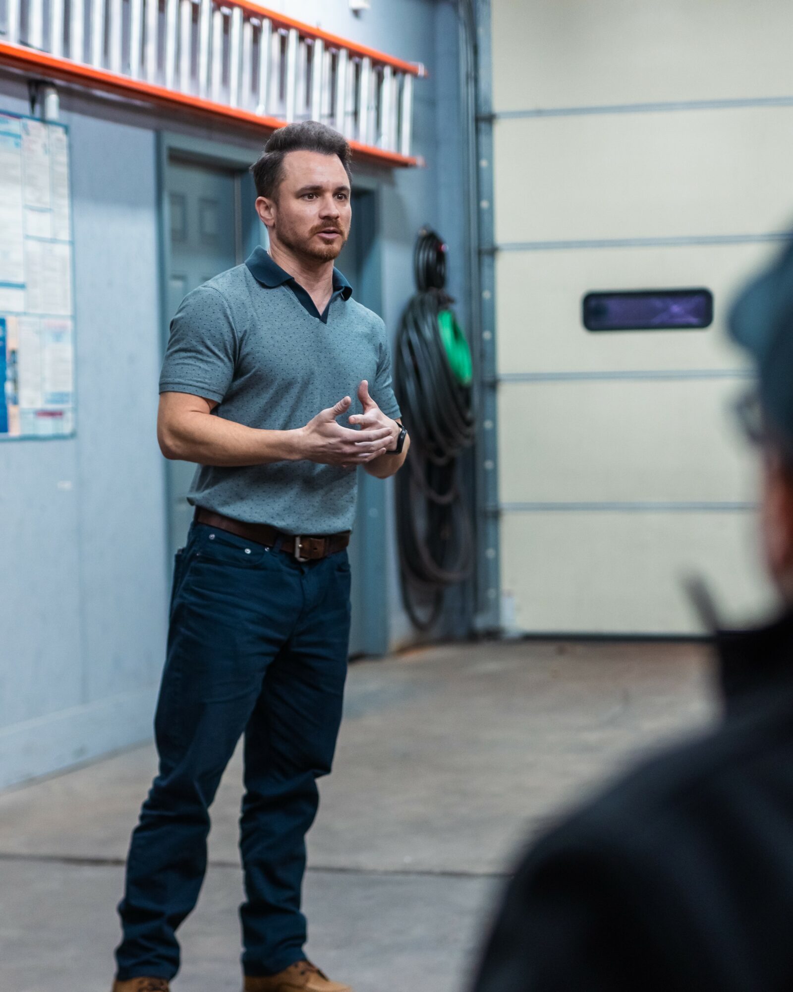 Man standing and speaking in an industrial setting with a garage door and equipment in the background.