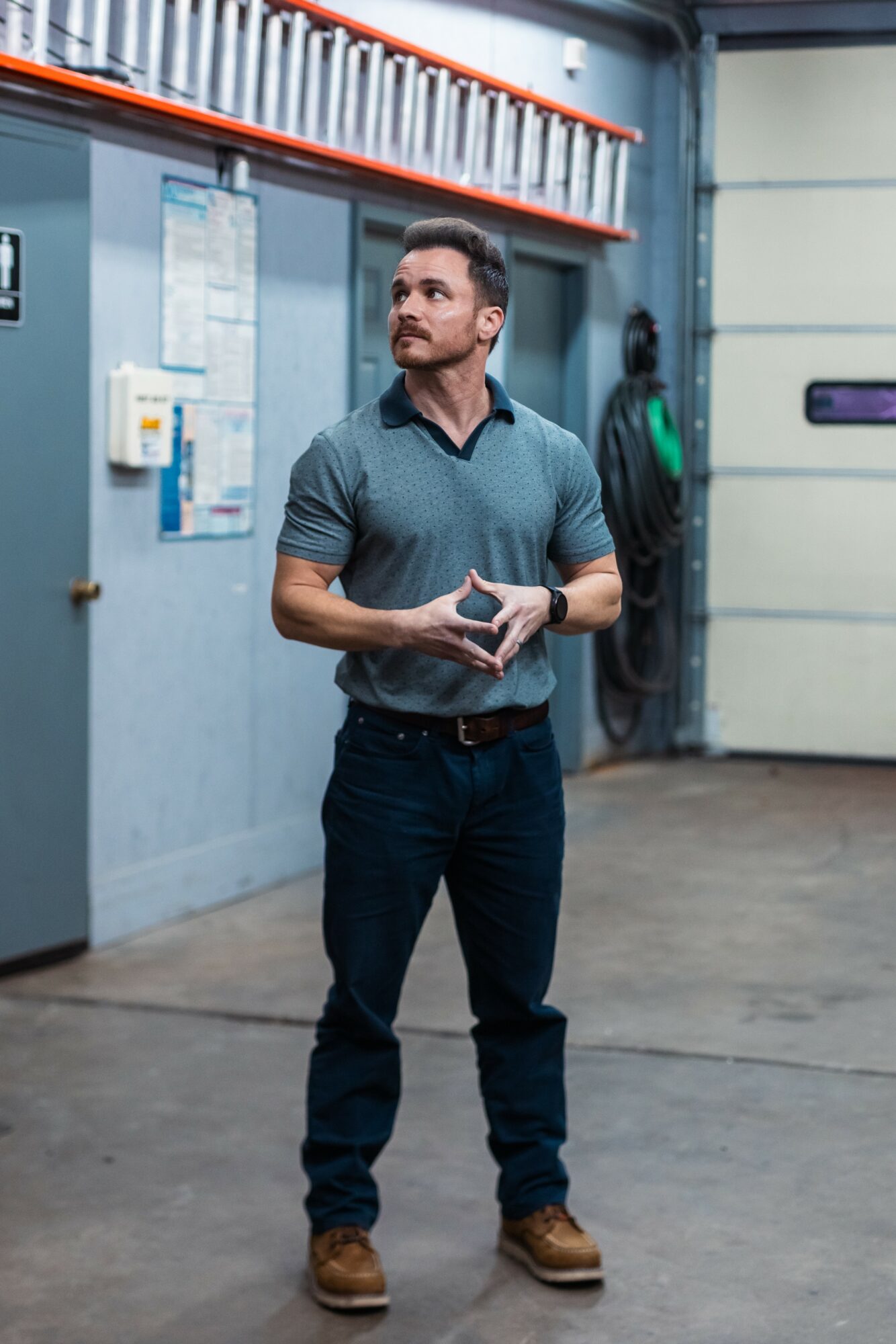 Man standing in industrial setting holding a phone, wearing a dark shirt and jeans, with a person in the background.