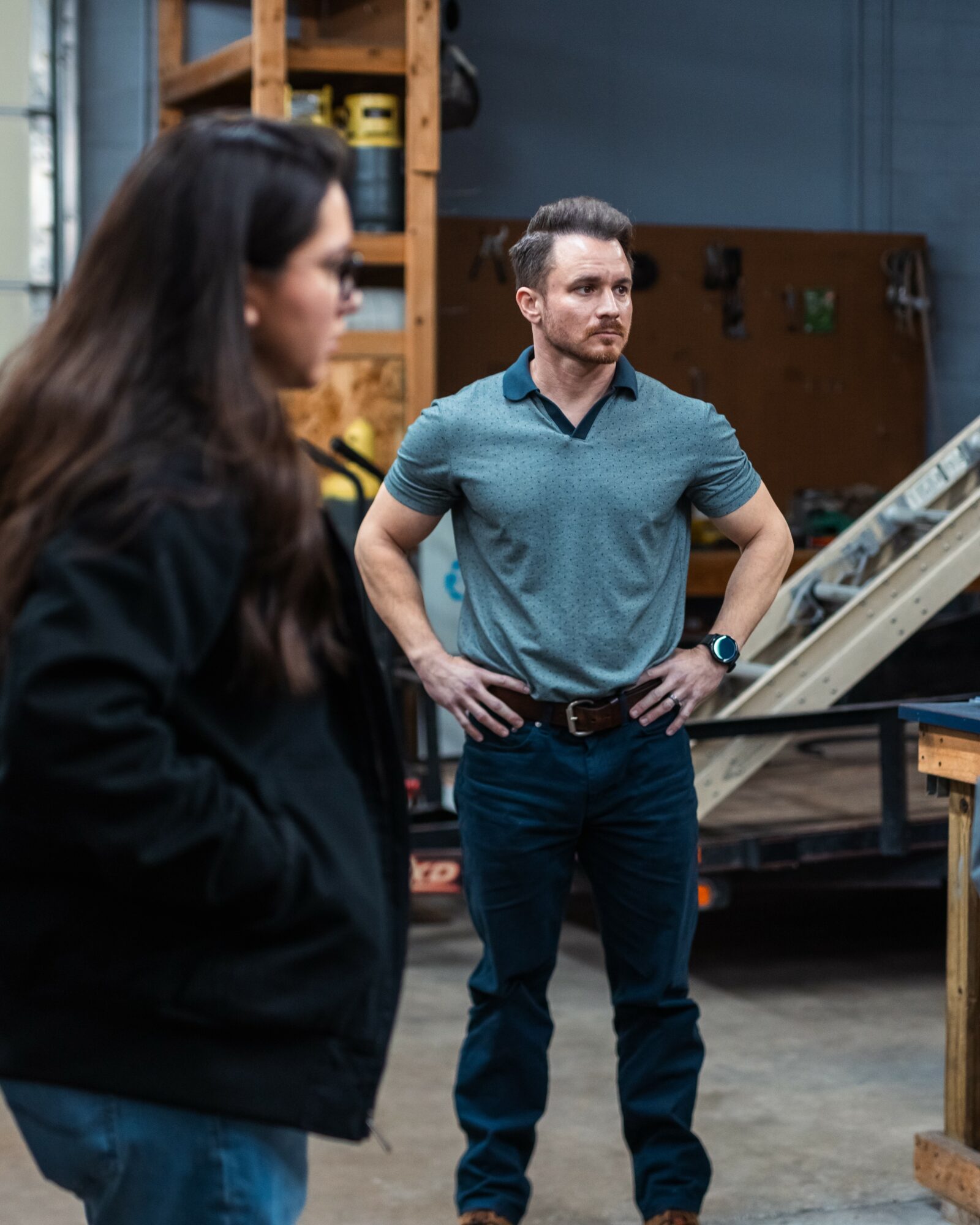 Man with hands on hips and woman with long dark hair in a workshop setting, shelves and equipment in background.