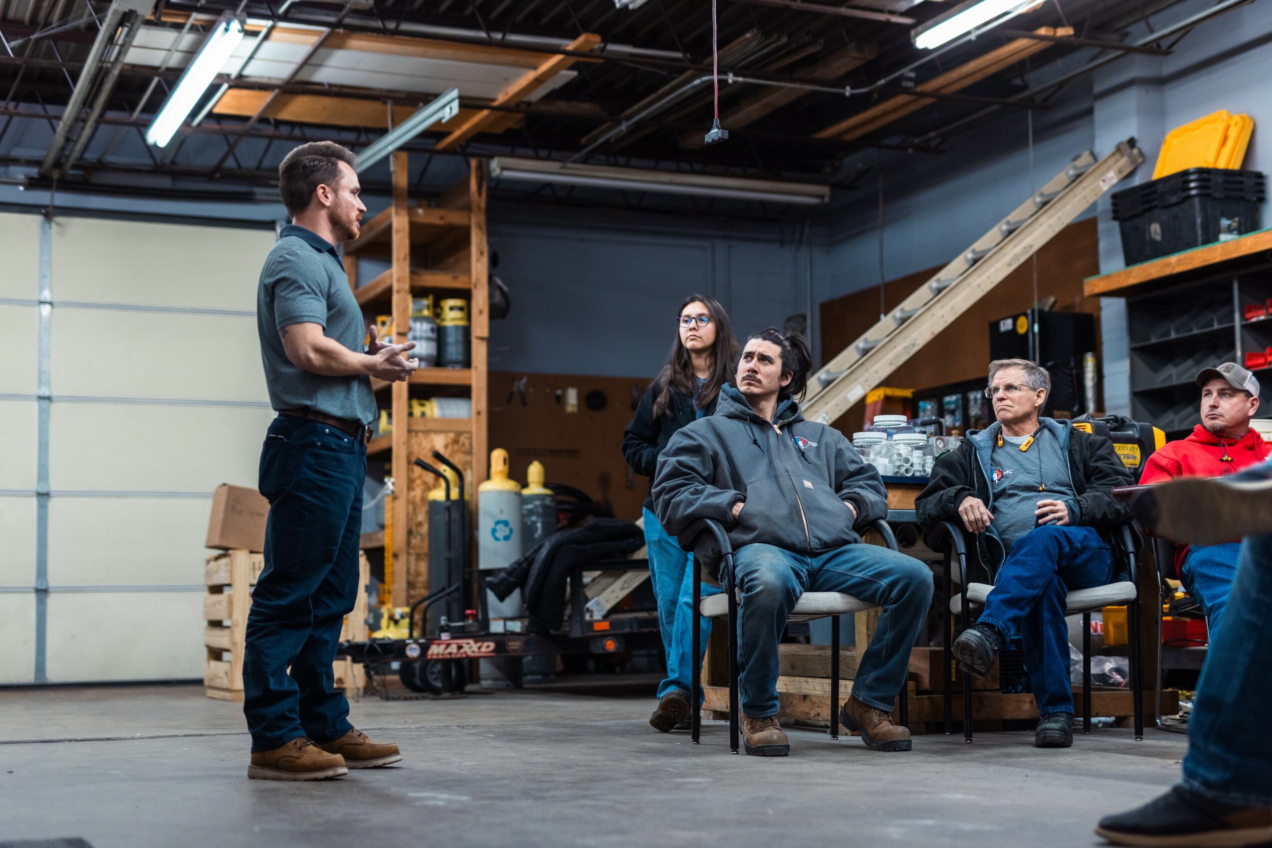 Four people in a workshop listening to a standing man, with shelves and equipment in the background.