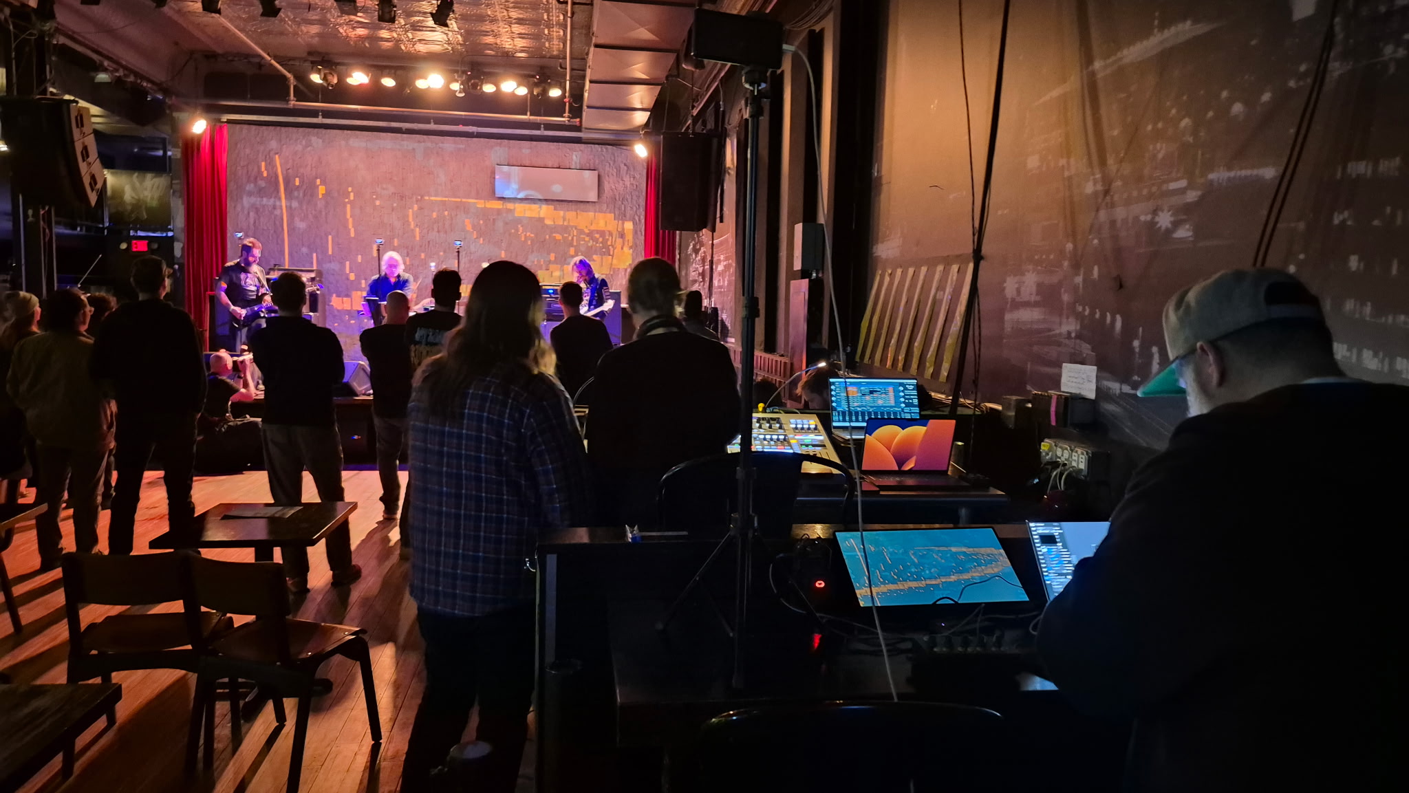 People watching a stage performance in a dimly lit venue with a sound engineer working at a mixing console.