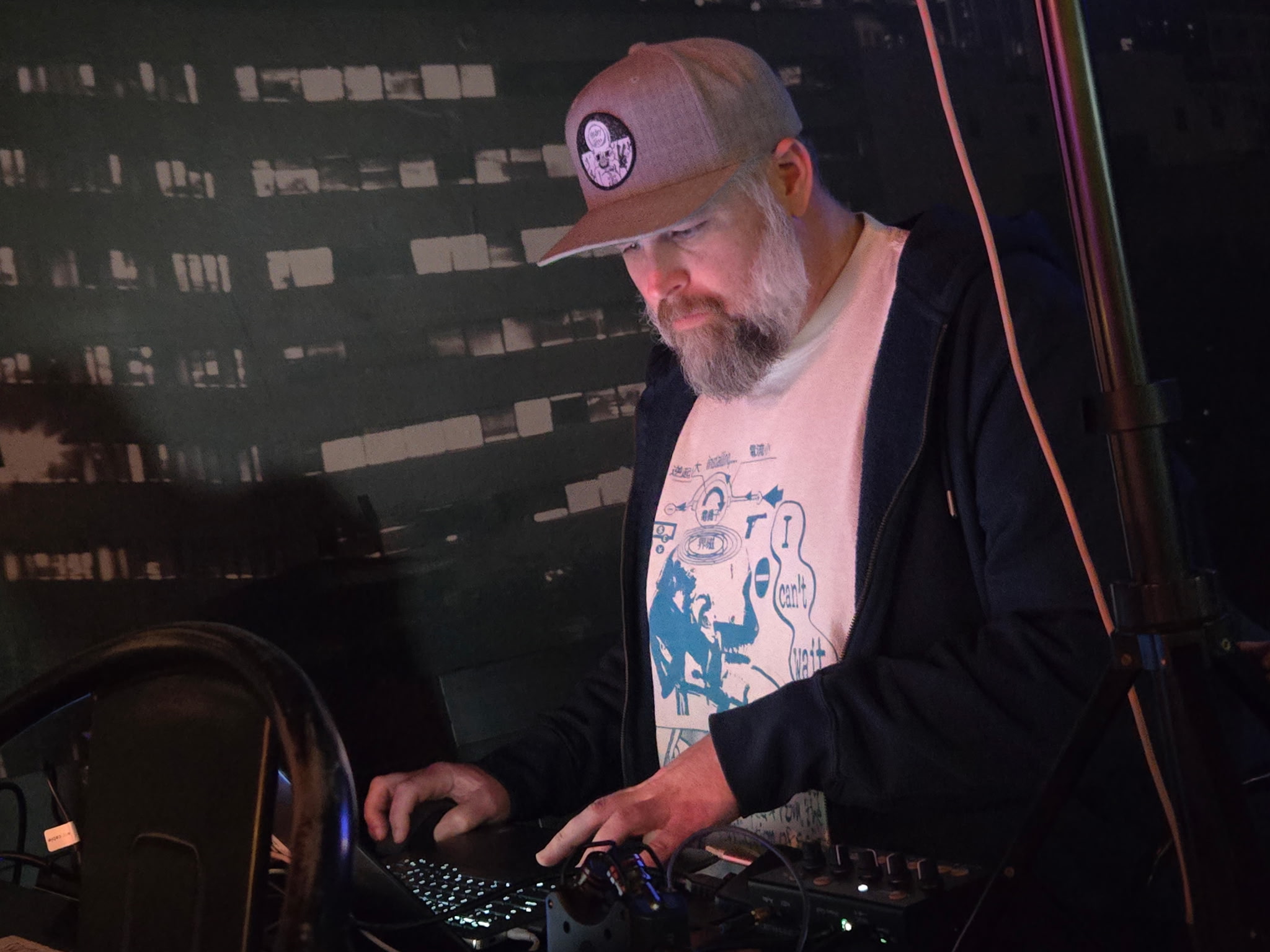 Man with a beard wearing a cap, jacket, and graphic t-shirt, operating DJ equipment at night, city lights in background.