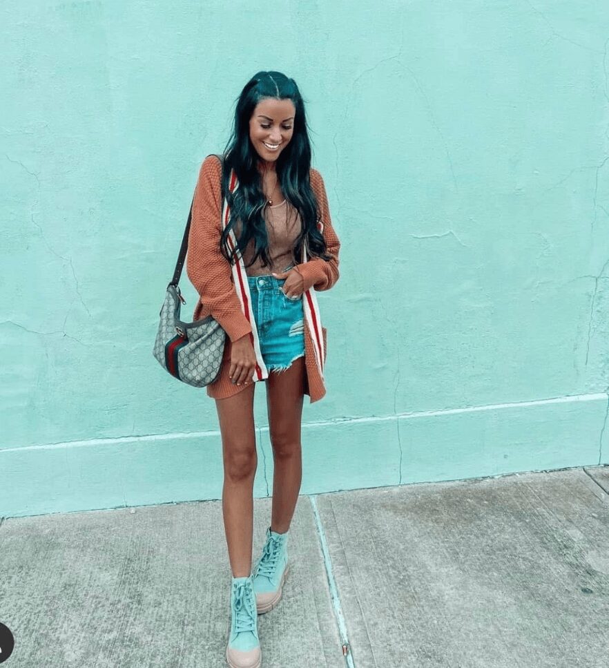 Young woman with long dark hair smiling, standing against a light blue wall, wearing a brown jacket, striped shorts, and light blue shoes.