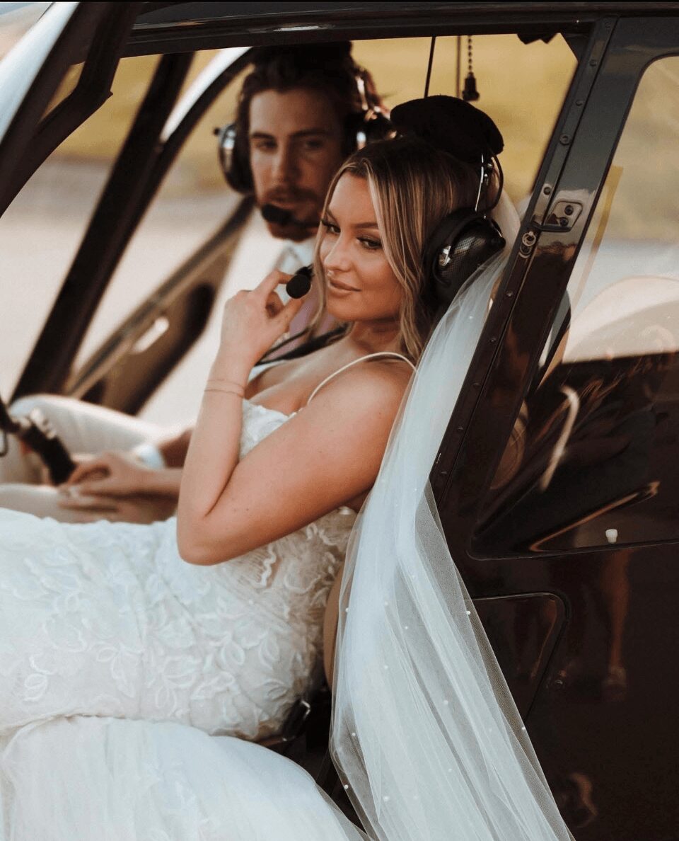 Bride in wedding dress sitting in helicopter, wearing headphones, smiling, with groom in background, during sunset.