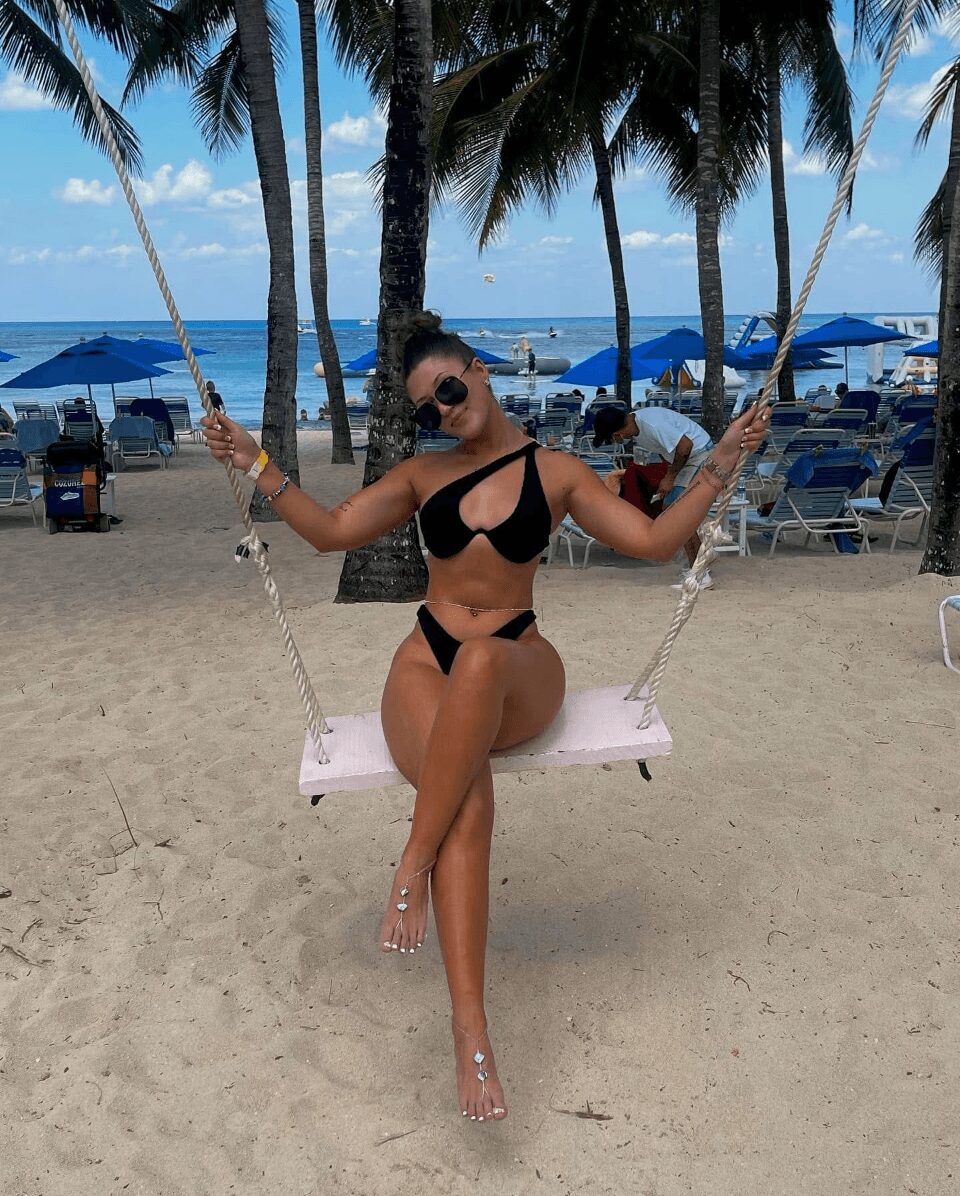 Woman in black bikini sitting on a swing at the beach, palm trees, umbrellas, and ocean in background.