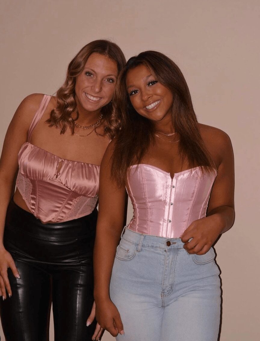 Two women smiling, one with wavy hair wearing a pink top and black pants, the other with straight hair in a pink corset and light jeans.
