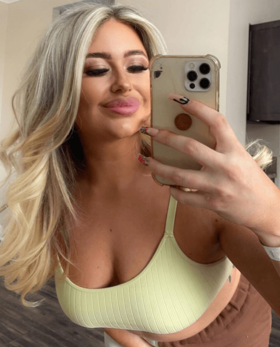 Woman with long blonde hair taking a selfie in a mirror, wearing a light green top, smiling with closed eyes.