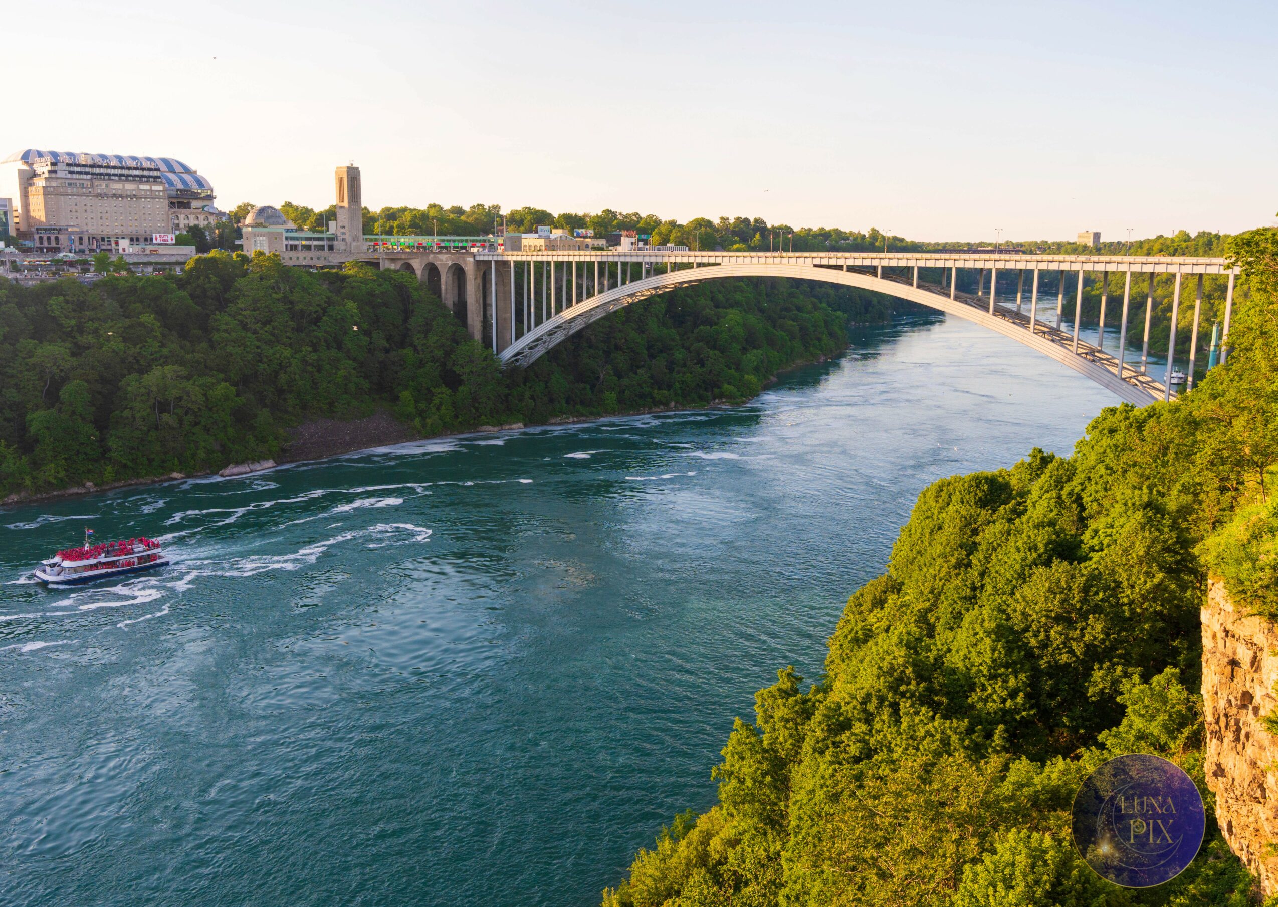 Bridge over a river with green trees on both sides, city buildings in the background, and a boat on the water.