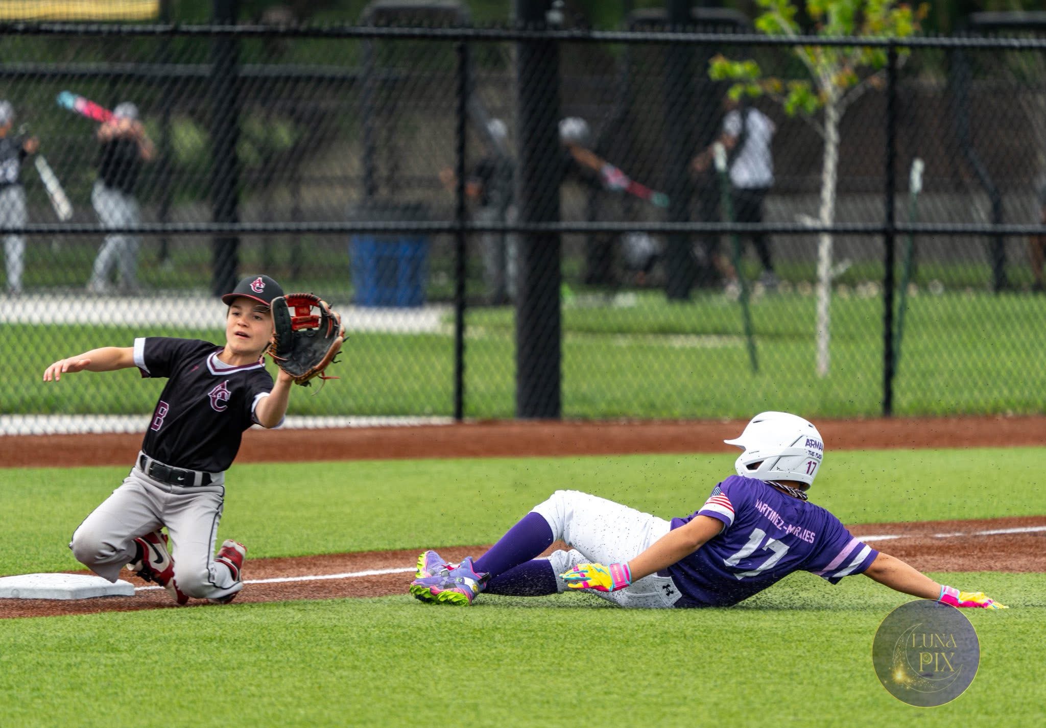 Baseball player sliding into base while fielder reaches with glove, on grassy field with fence and spectators in background.