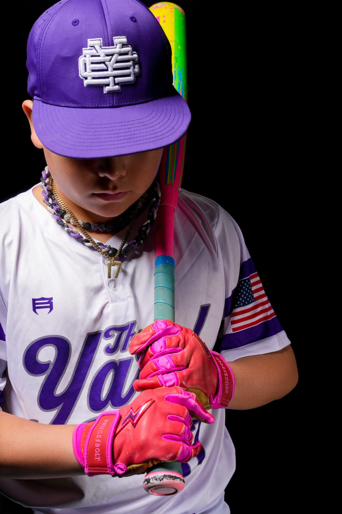 Child wearing a purple cap, white jersey, pink gloves, holding a baseball bat, against a black background.
