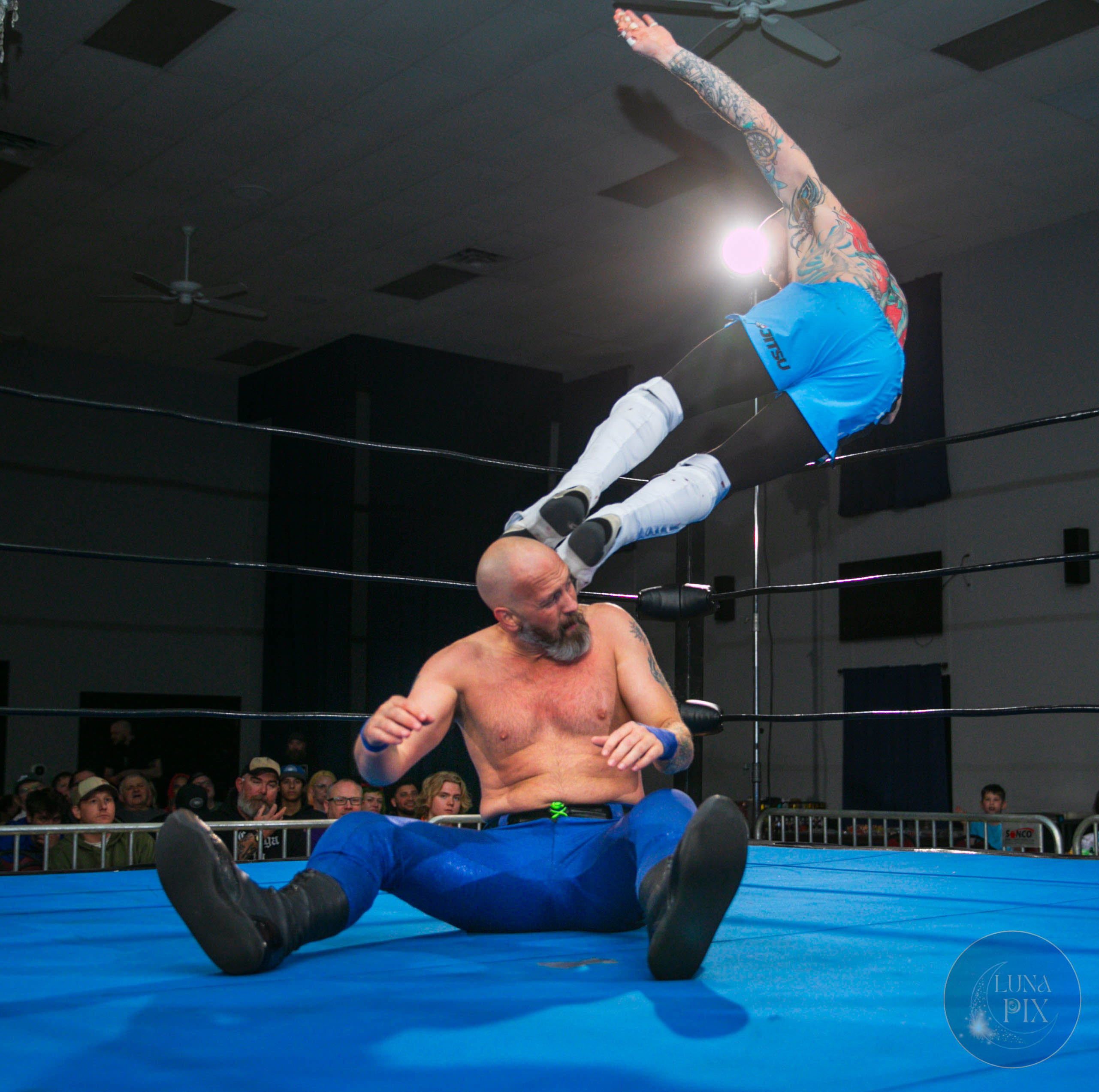 A wrestler with tattoos is mid-air above another wrestler sitting on the ring floor, in a wrestling match.