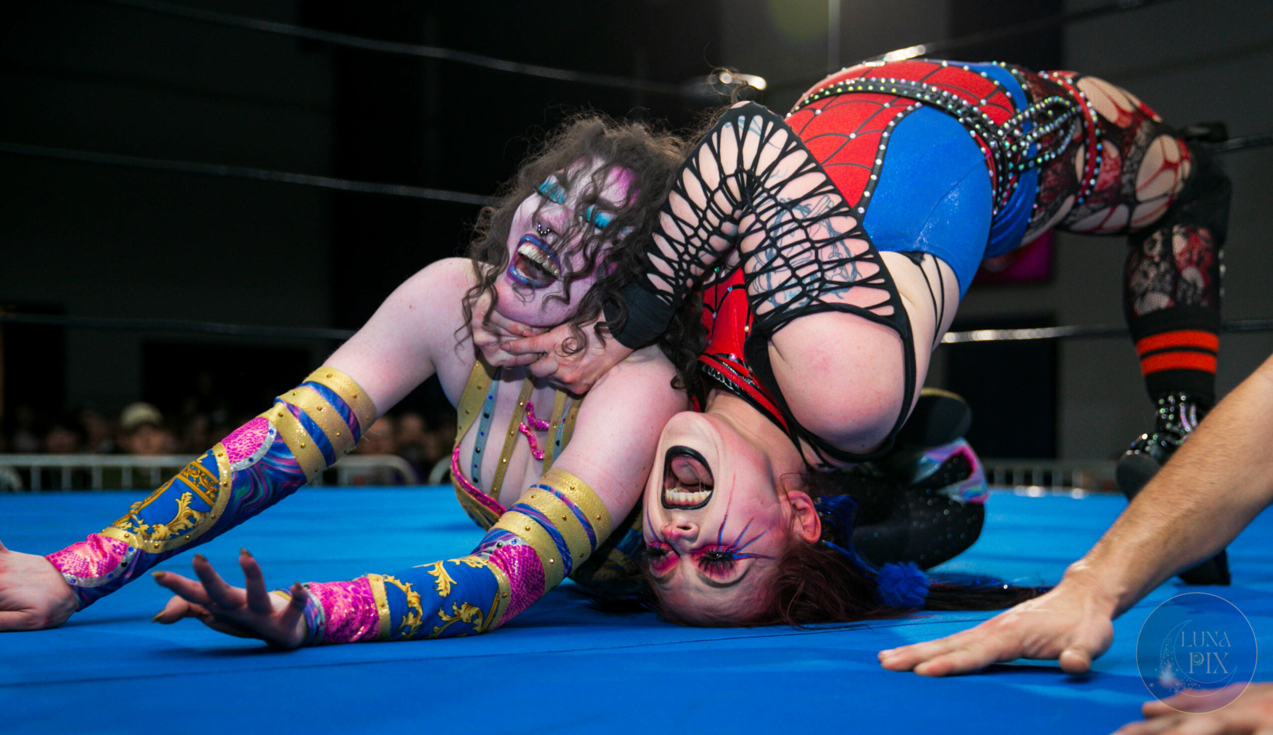Two female wrestlers in colorful costumes engage in a match on the mat, one on top of the other, in a wrestling ring.